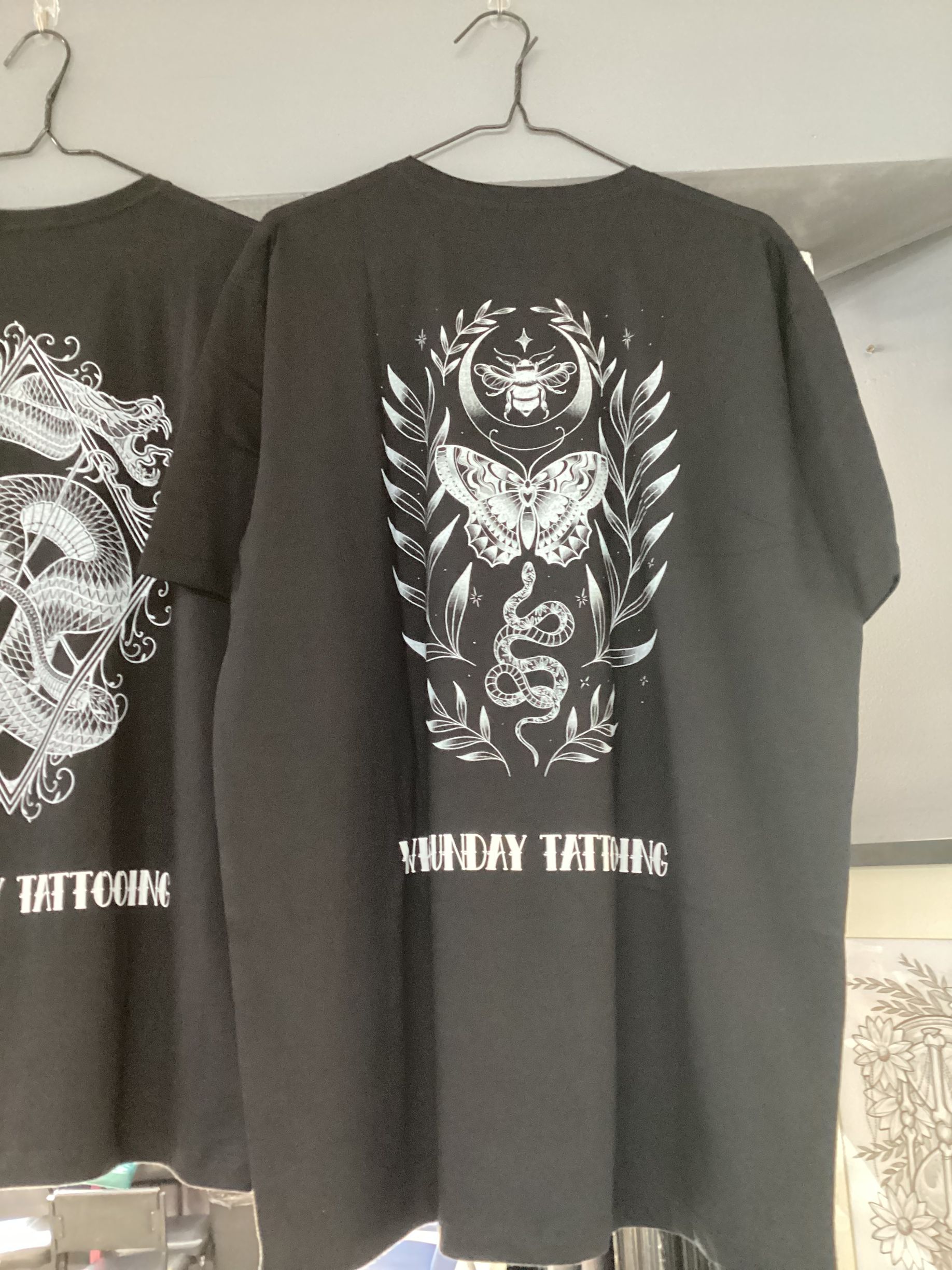 Two Black T-Shirts with Silver Graphic Designs Hanging on A Rack — Whitsunday Tattooing Airlie Beach in Whitsundays, QLD