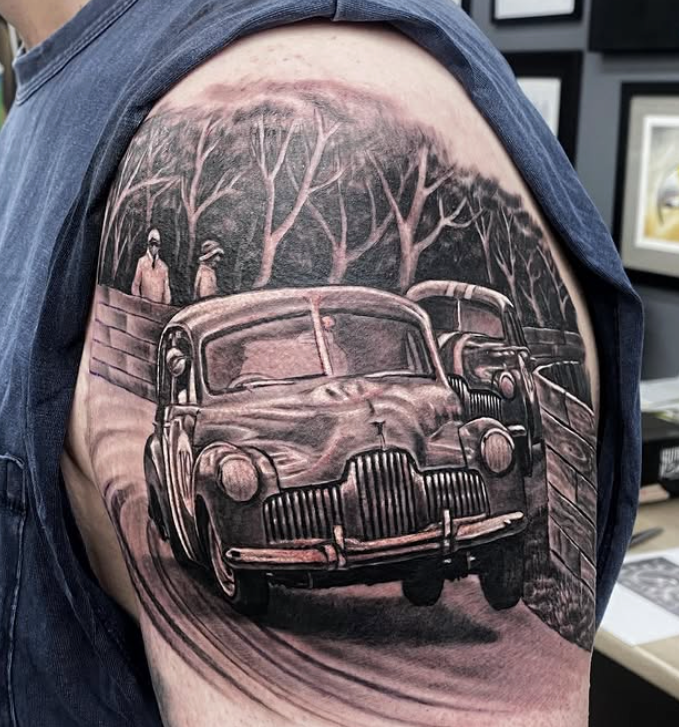 Black and gray tattoo of a vintage car driving on a road, trees in background, on a person's shoulder. — Whitsunday Tattooing Airlie Beach in Whitsundays, QLD