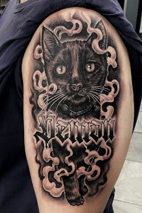 Black cat tattoo on an arm, with stylized name, framed by smoky clouds.— Whitsunday Tattooing Airlie Beach in Proserpine, QLD