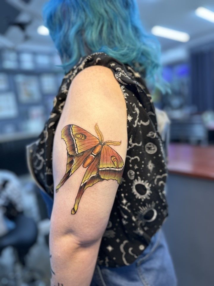 A person with blue hair and a moth tattoo on their upper arm. They are wearing a black patterned vest. — Whitsunday Tattooing Airlie Beach in Whitsundays, QLD