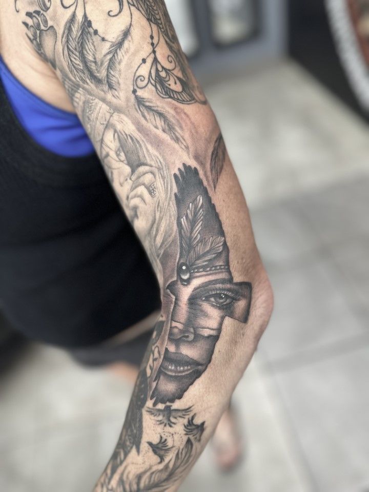 Arm tattoo with a face and feathers, in grayscale. — Whitsunday Tattooing Airlie Beach in Whitsundays, QLD