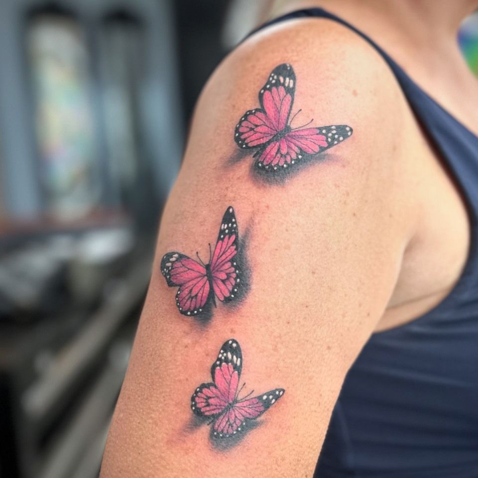 Three pink butterfly tattoos on a person's upper arm, with black detailing and shading. — Whitsunday Tattooing Airlie Beach in Whitsundays, QLD
