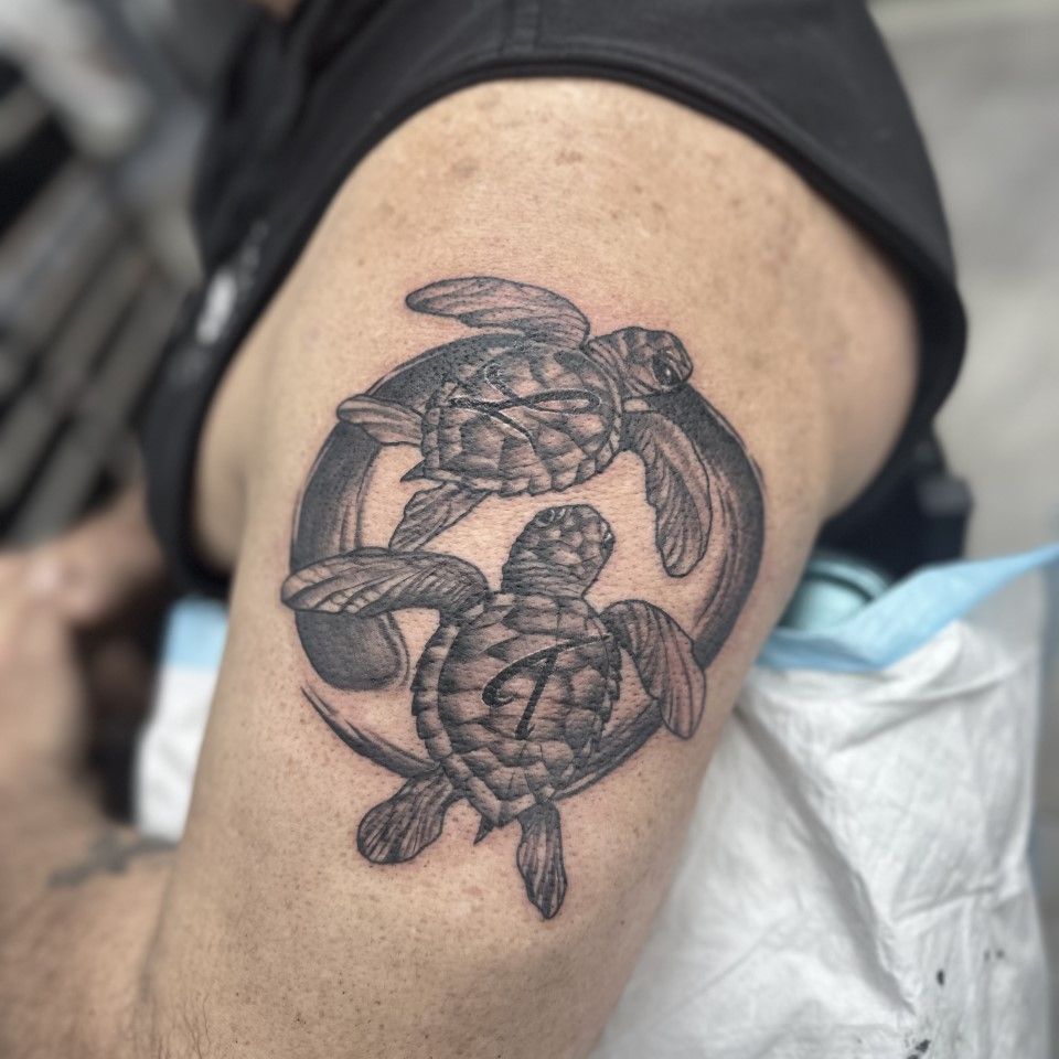 Tattoo of two sea turtles swimming through a ring, inked in black on a person's upper arm. — Whitsunday Tattooing Airlie Beach in Whitsundays, QLD