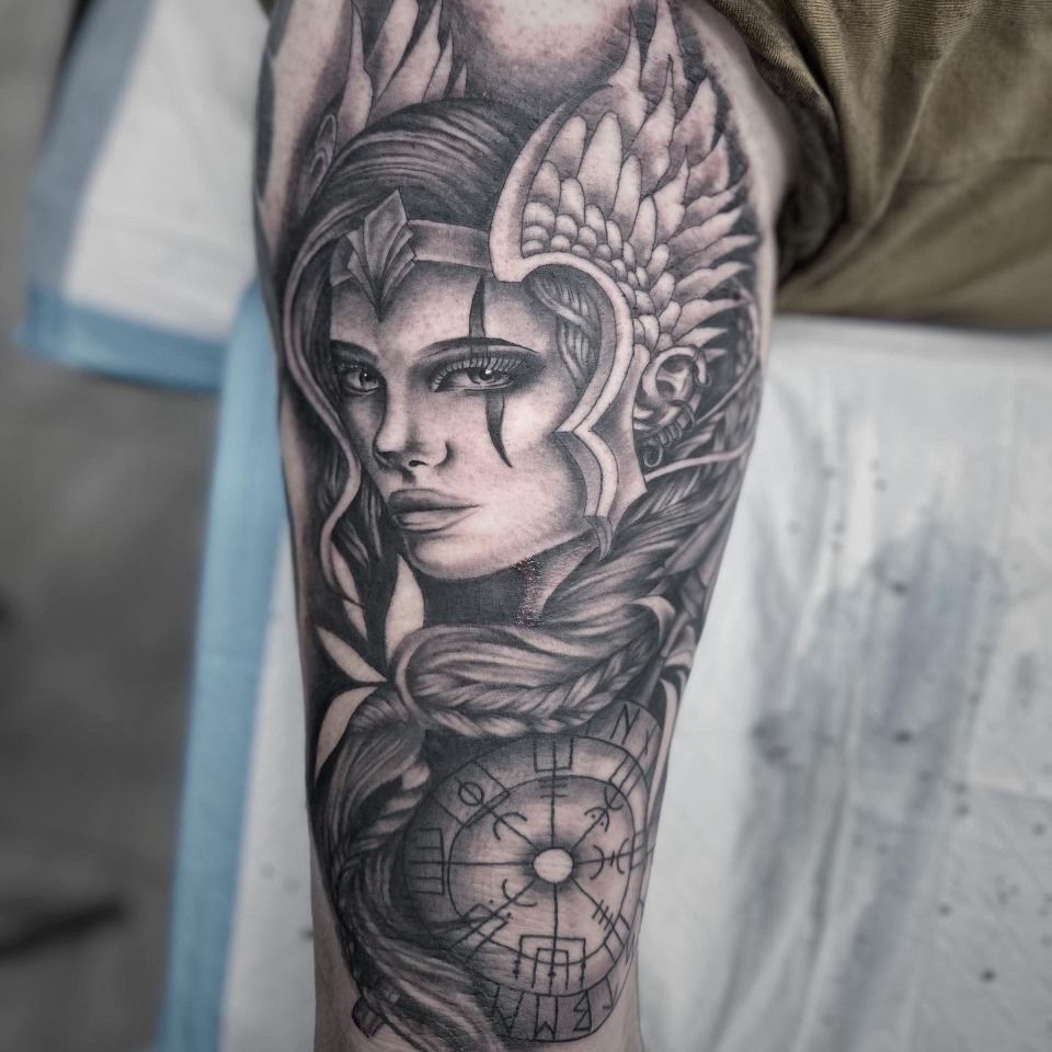 Black and gray tattoo of a woman with a winged headdress and a compass, on an arm.