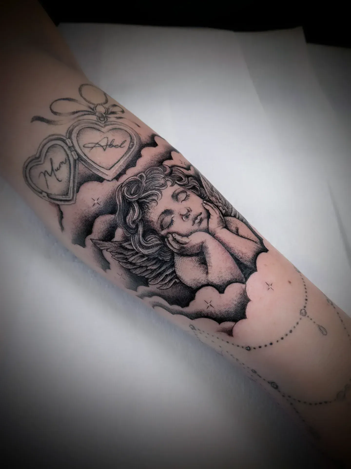 Tattoo of a sleeping cherub in clouds on an arm, with two heart lockets and a beaded chain.