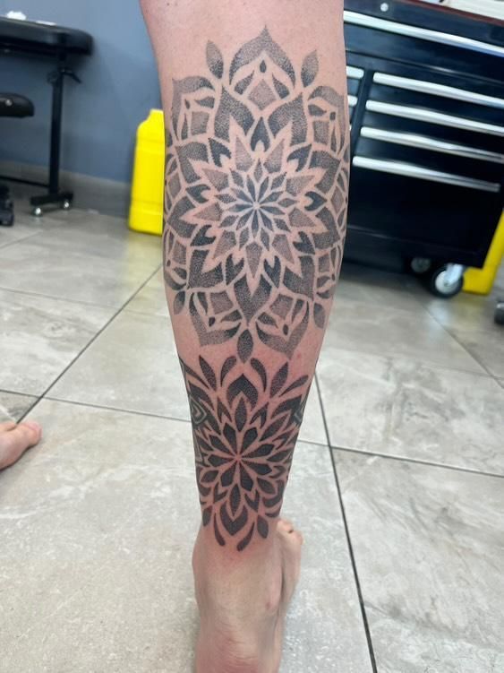 Black mandala tattoo on a leg, with detailed geometric patterns. Tattoo spans the calf to ankle.