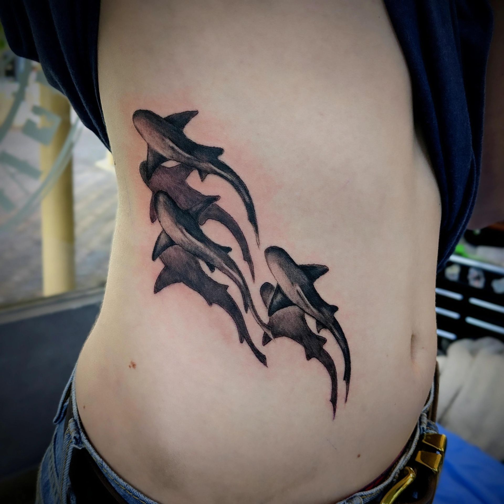 Black shark tattoo on a person's side, possibly torso. Sharks in a vertical cluster. Person wears blue shirt, jeans.