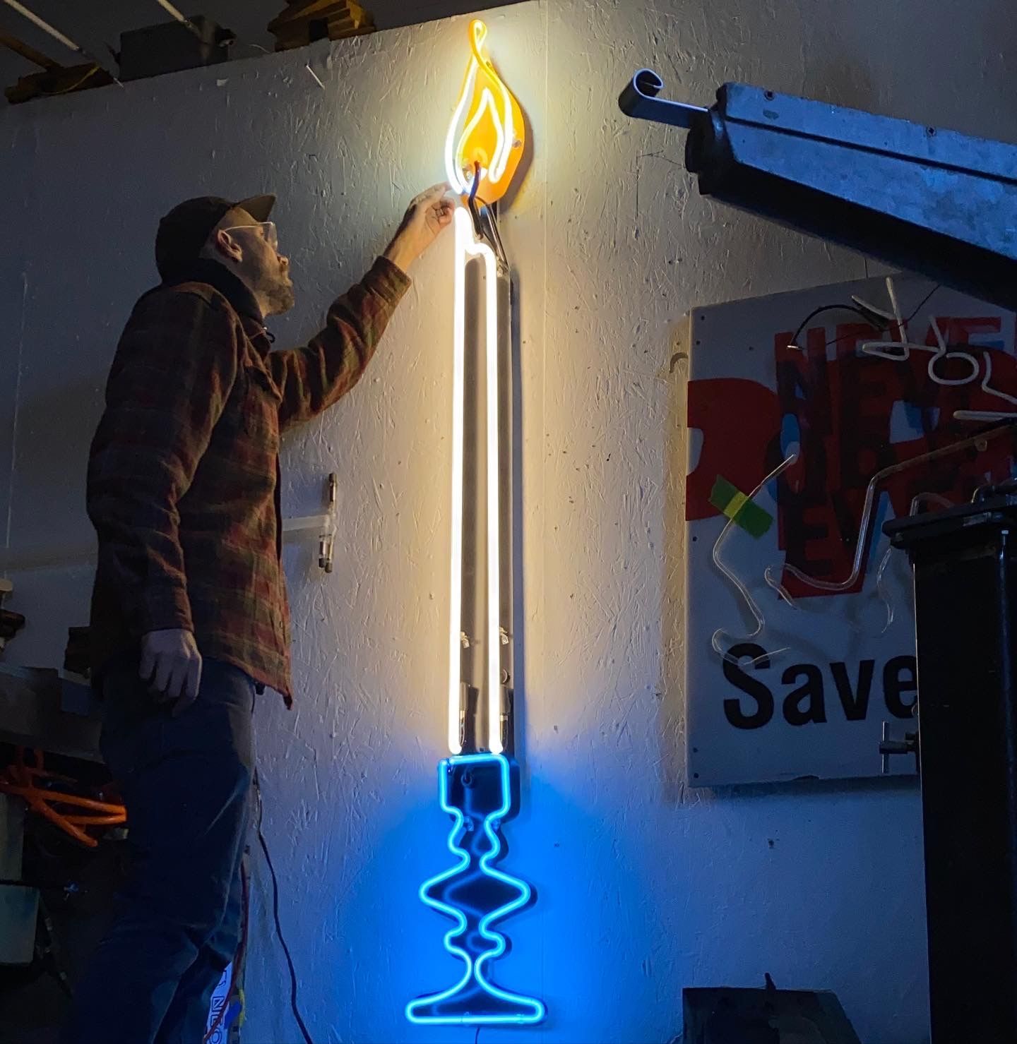 Man working on a neon candle. Yellow flame, white shaft, blue base. Inside workshop.