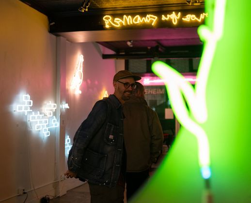 Artist Yale Wolf, in a cap and denim jacket walks through a dimly lit gallery decorated with colorful neon art installations.