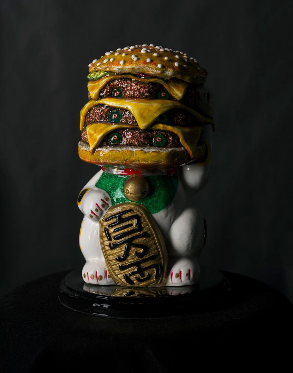 A ceramic beckoning cat figurine with a stack of hamburgers for a head, set against a dark background.