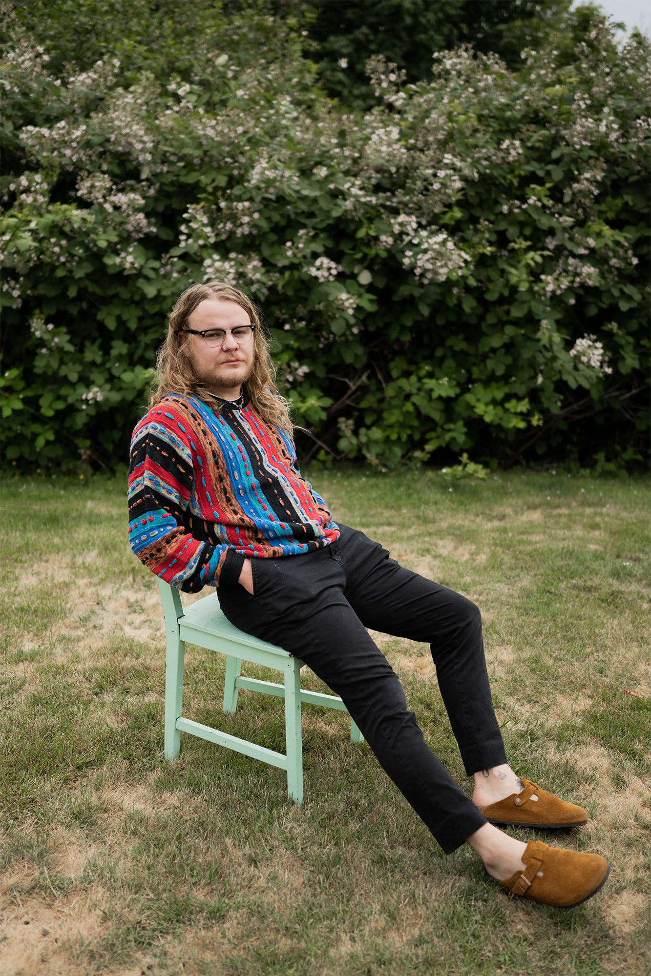 Jackson O'Rourke with long hair, glasses, colorful sweater, black pants, brown shoes, sitting on green chair in yard.