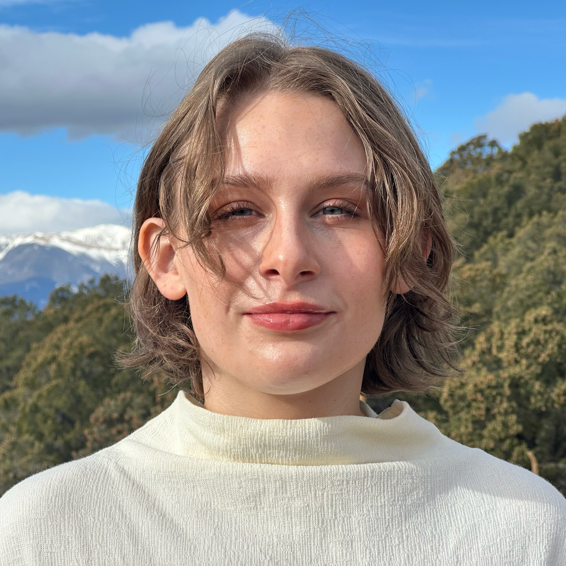 Woman with short brown hair, wearing a cream turtleneck, smiles in front of a mountain and cloudy sky.