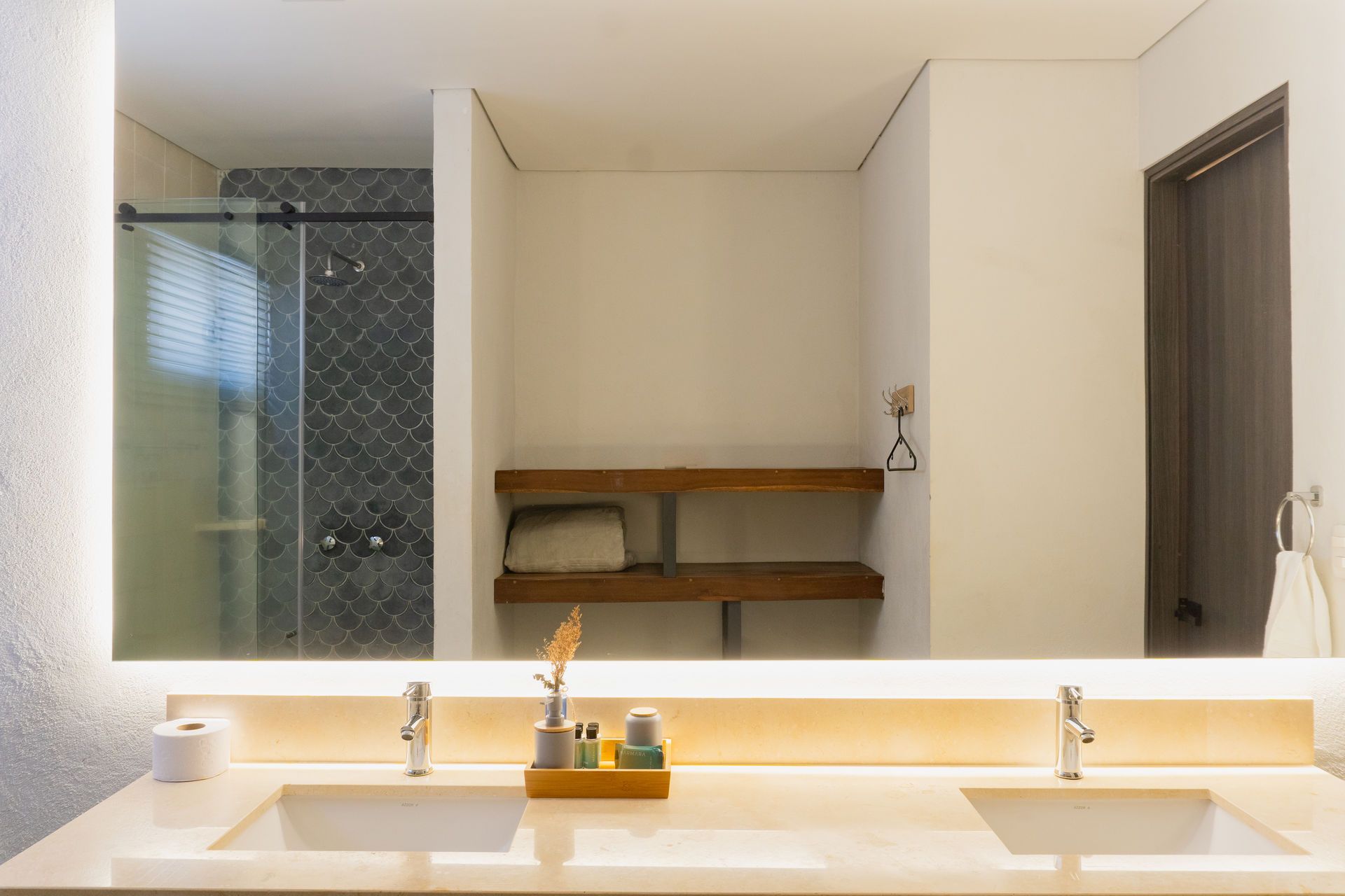 Baño con un gran espejo iluminado que refleja la ducha y los lavabos. Encimera y estanterías color crema.