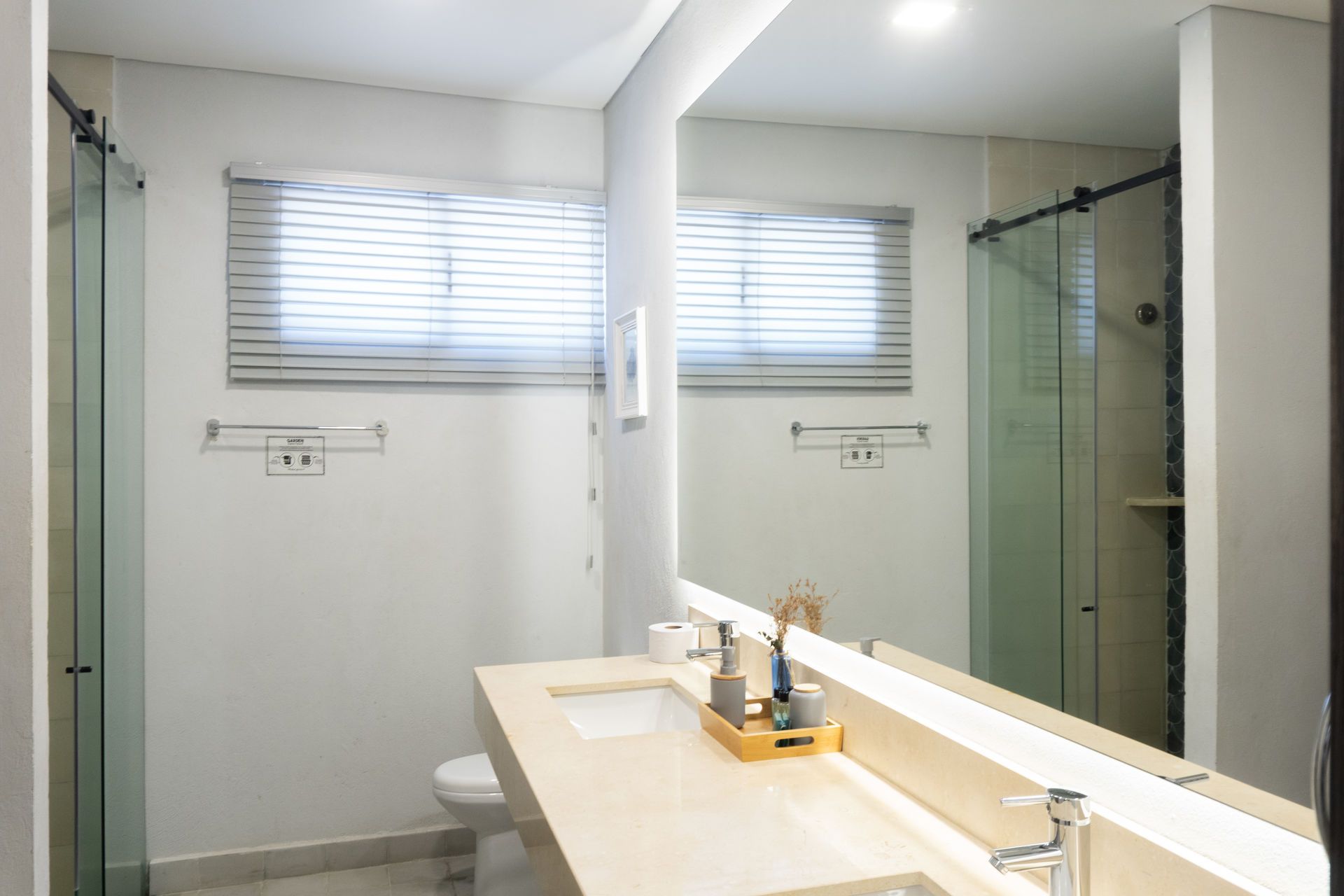 Baño con gran espejo, lavabo doble, ducha y ventana con persianas.