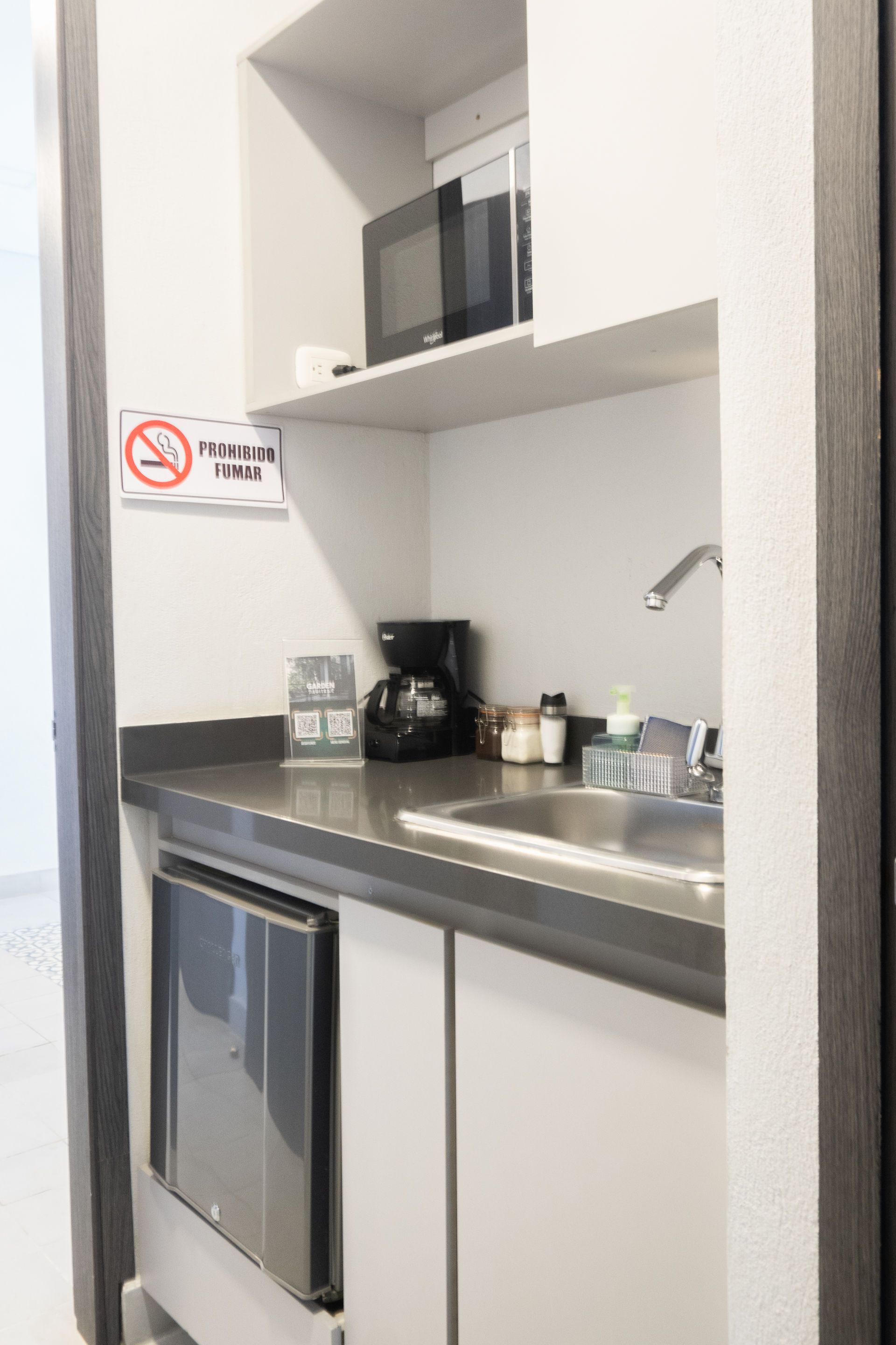Pequeña cocina con microondas, cafetera, fregadero, mininevera y cartel de 