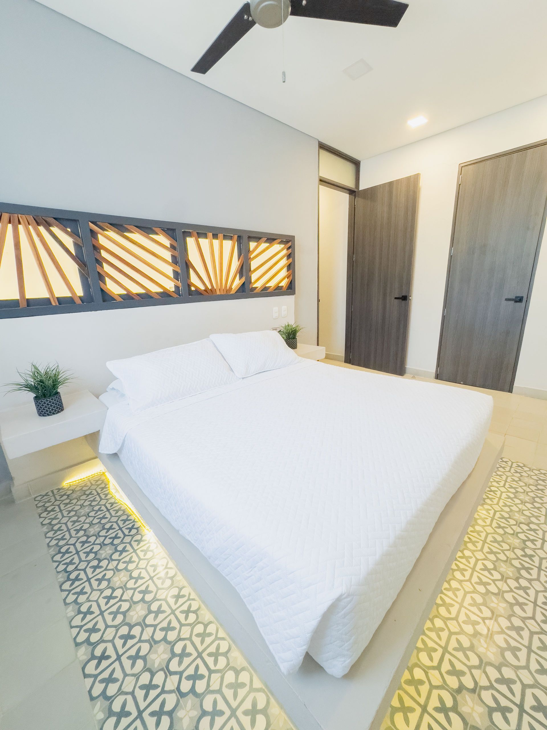 Dormitorio con cama blanca, alfombra estampada y cabecero de madera con motivos geométricos.