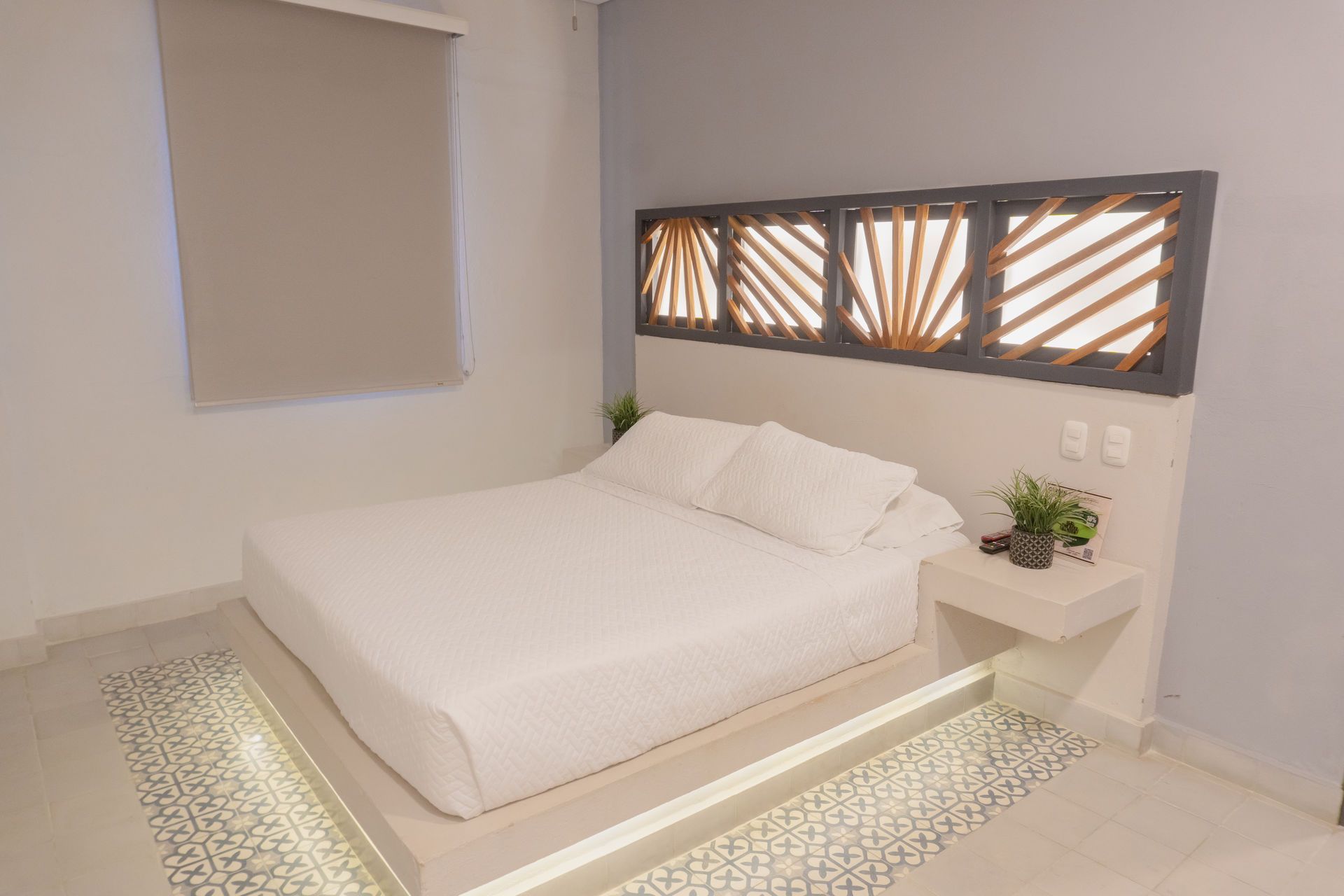 Dormitorio con cama, mesita de noche y cabecero decorativo. Paredes grises, ropa de cama blanca y suelo de baldosas.