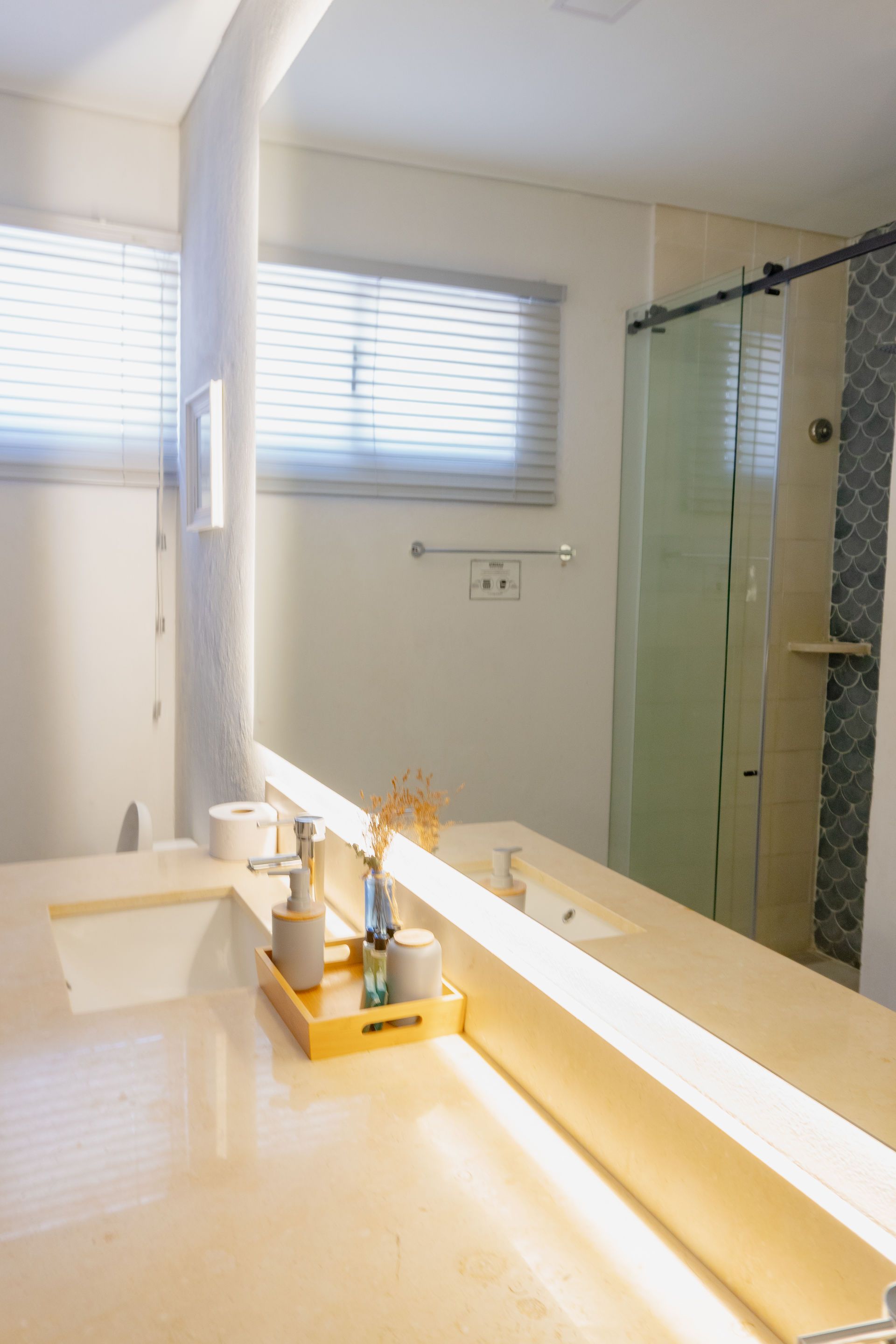 Baño con gran espejo que refleja un tocador, ducha y ventana con persianas.