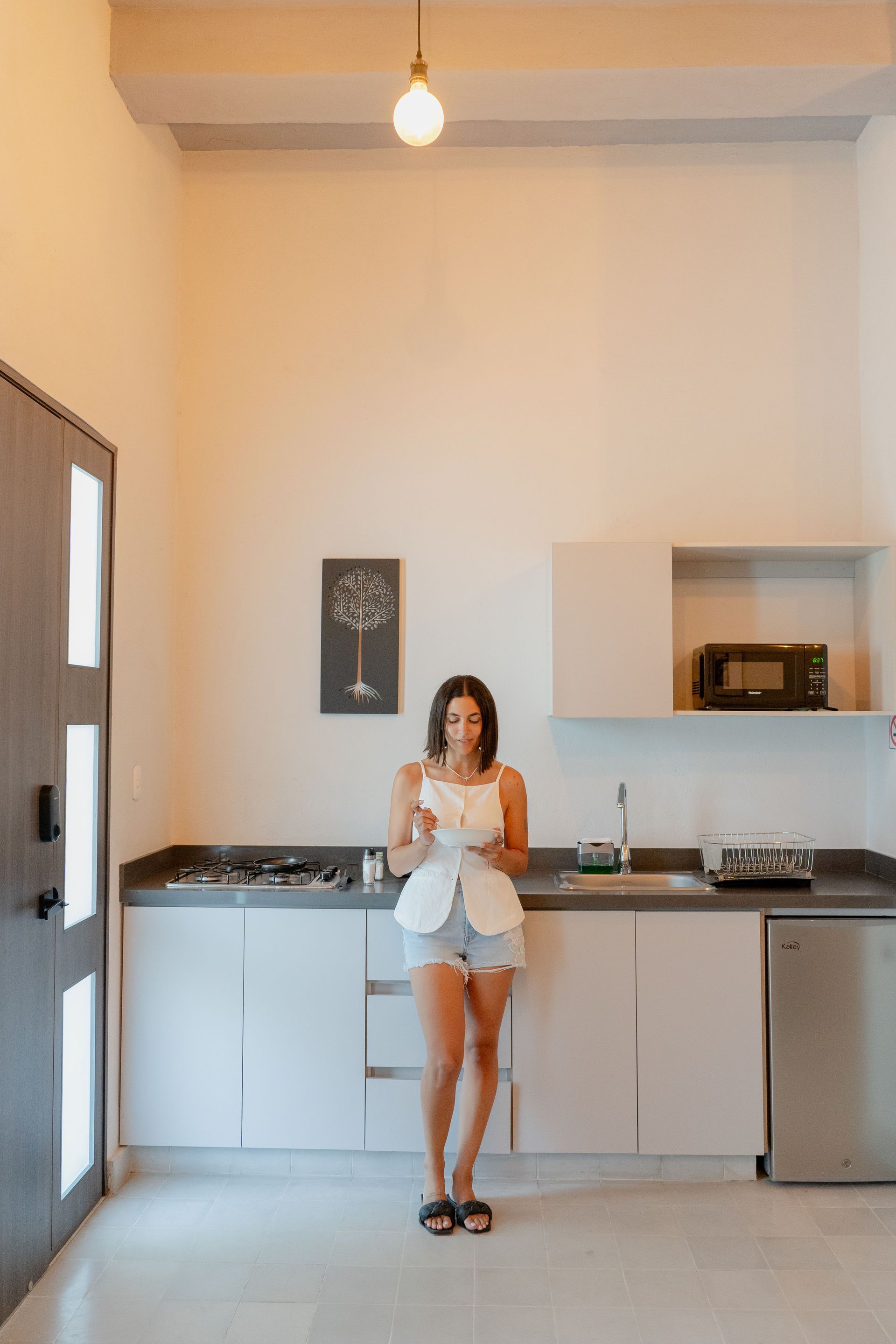 Mujer en pantalones cortos en una cocina moderna, sosteniendo una taza. Habitación luminosa de diseño minimalista.