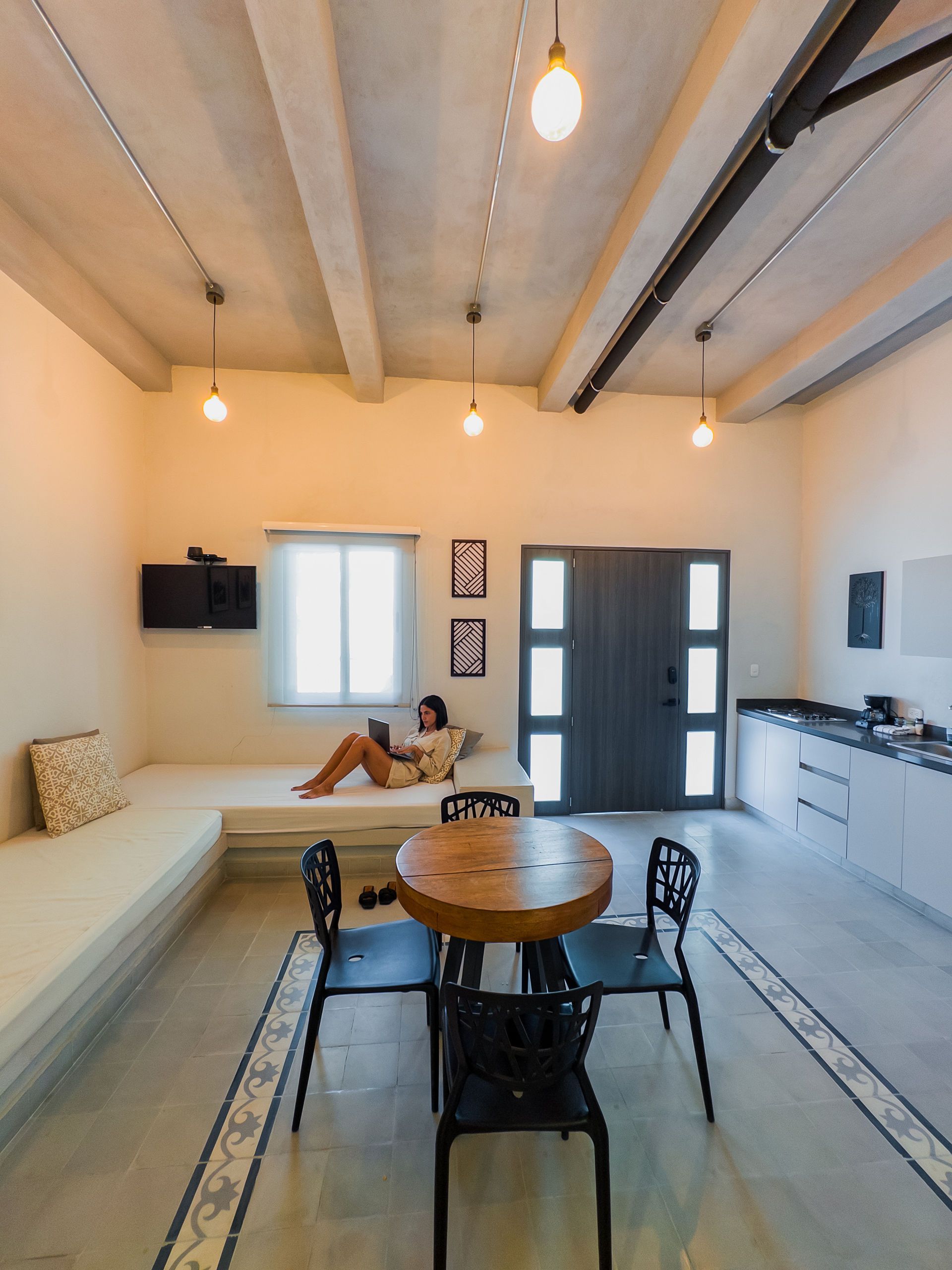 Una persona se relaja en un banco empotrado en una habitación moderna, con una tableta en la mano. Cerca hay una mesa con sillas y una puerta abierta.