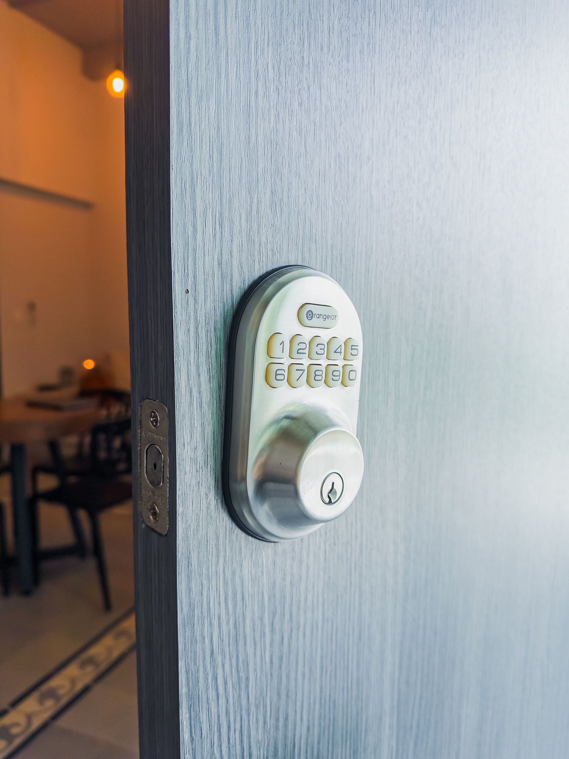 Cerradura con teclado plateado en una puerta gris. Interior de la habitación visible al fondo.