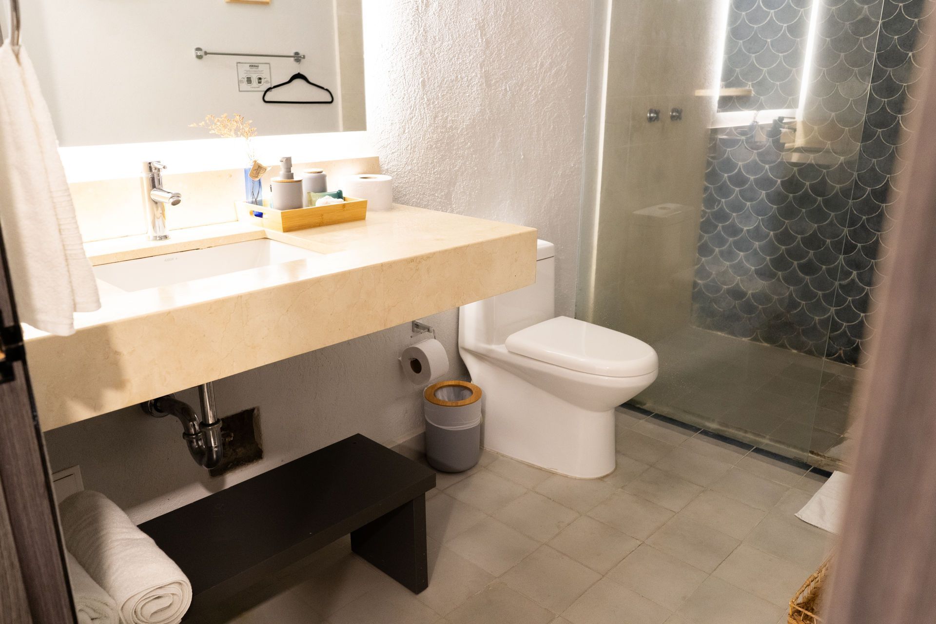 Baño con lavabo, inodoro y ducha. Hay un pequeño banco debajo del lavabo.