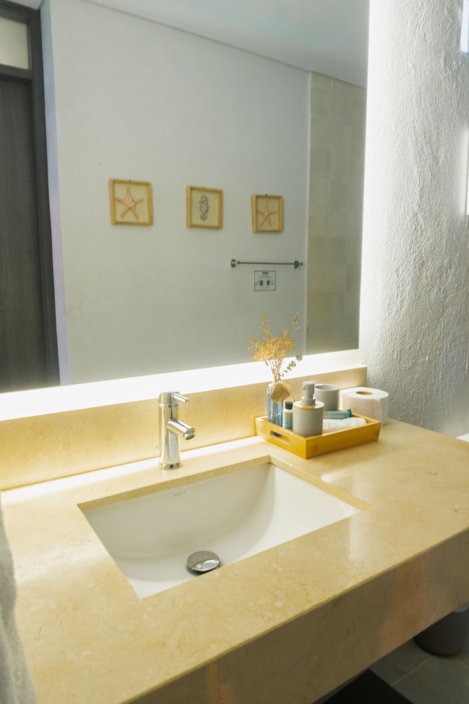 Baño con lavabo blanco, encimera beige, espejo grande y arte de conchas marinas.