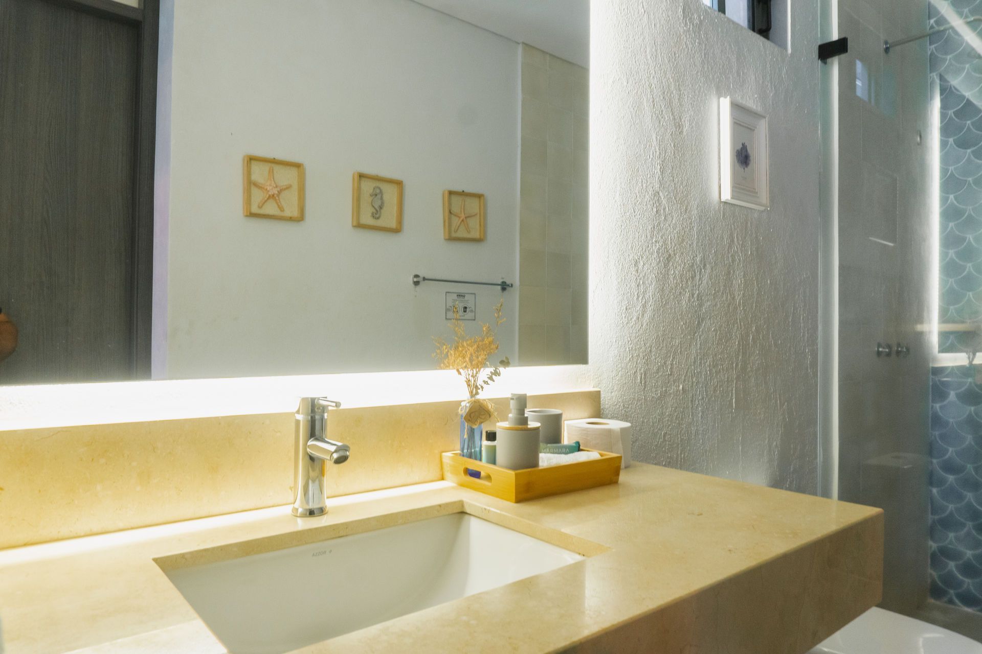 Baño con encimera beige, lavabo rectangular y espejo grande. Una obra de arte decorativa con estrellas de mar cuelga sobre el lavabo.