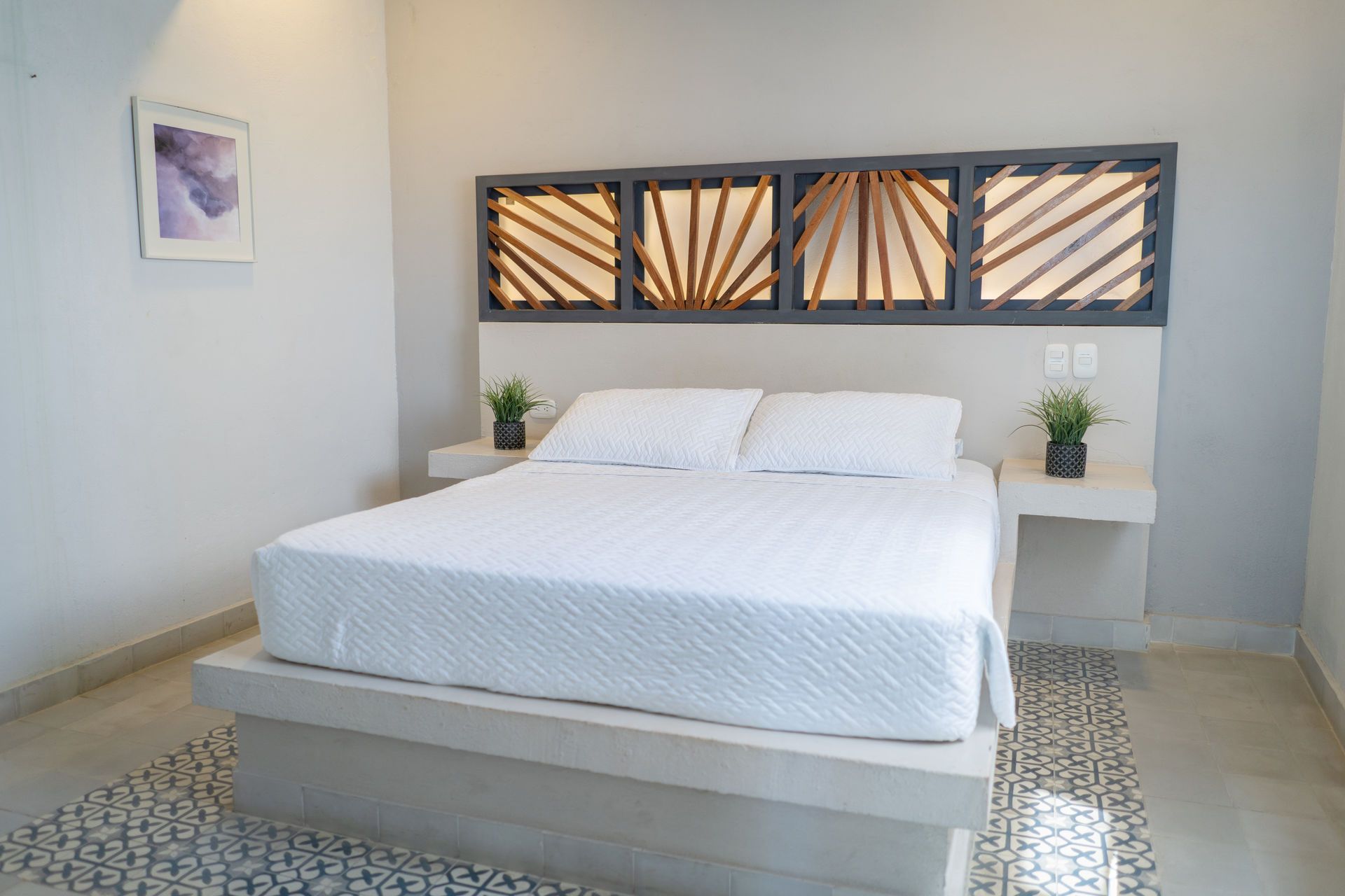 Dormitorio con cama, ropa de cama blanca, cabecero empotrado y mesitas de noche, obras de arte y suelo de baldosas estampadas.