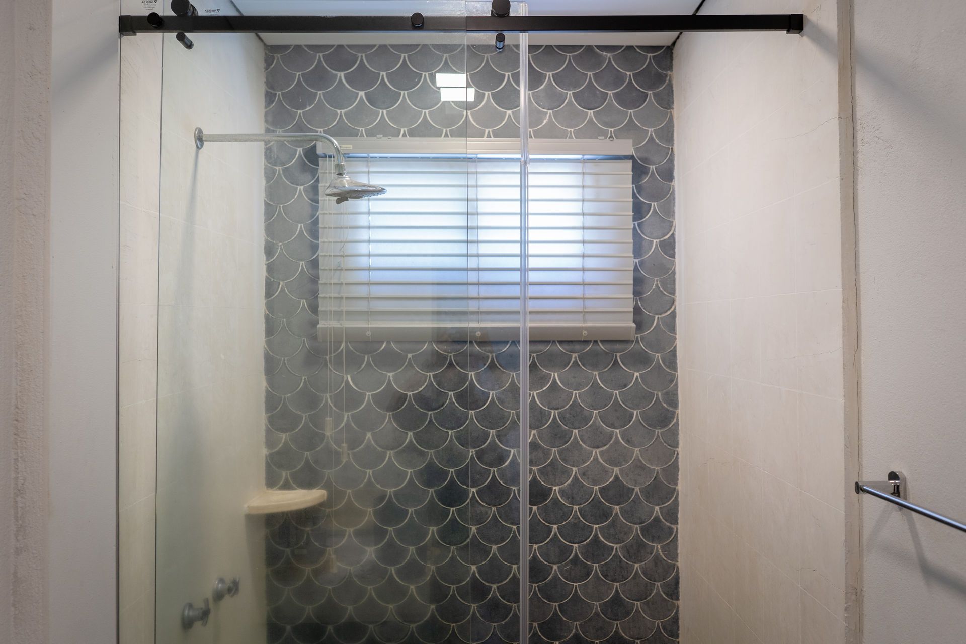 Cabina de ducha con puertas de cristal, pared de azulejos estampados, ventana con persianas y toallero.