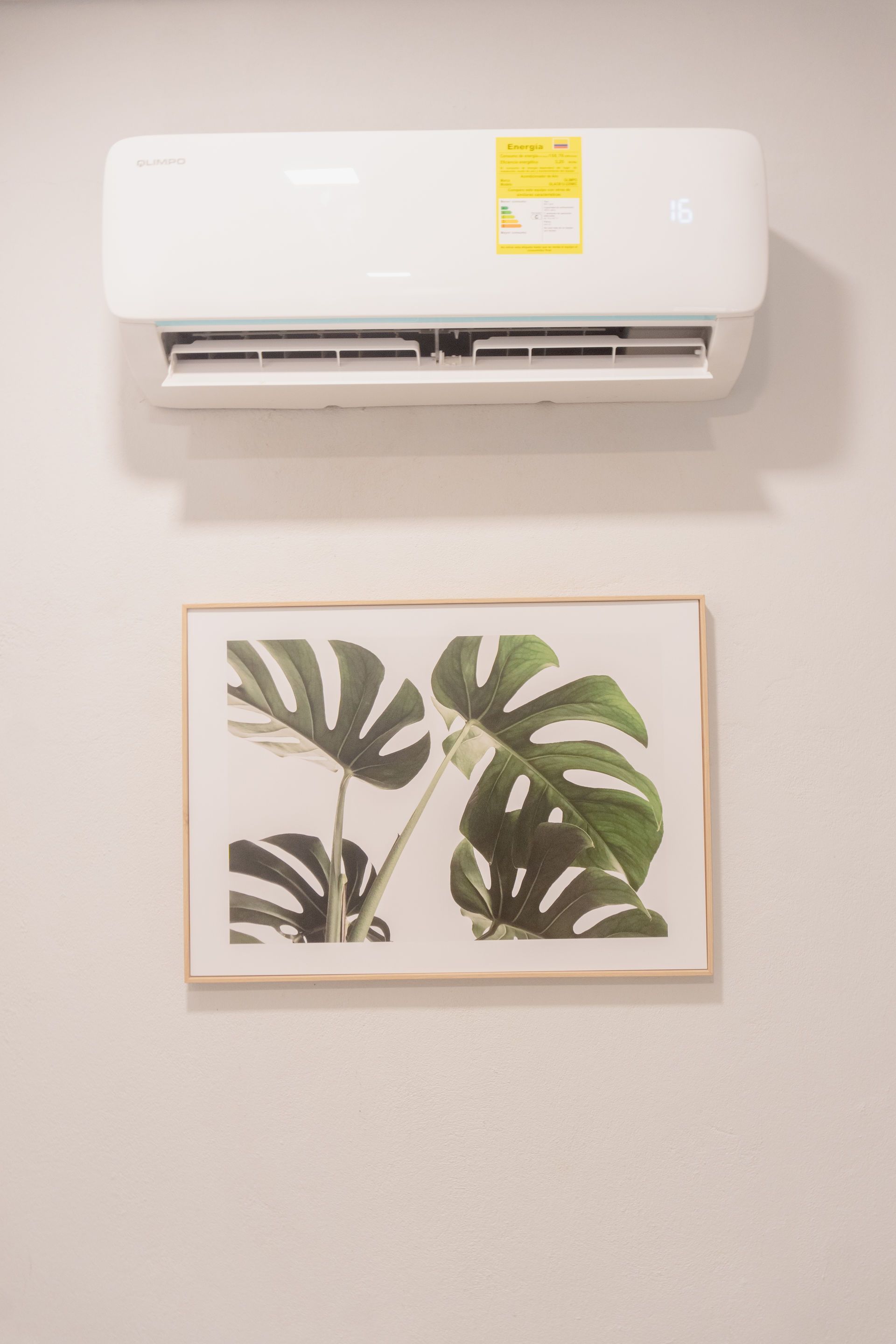 Aire acondicionado blanco sobre un cuadro impreso de hojas de Monstera en una pared de color claro.