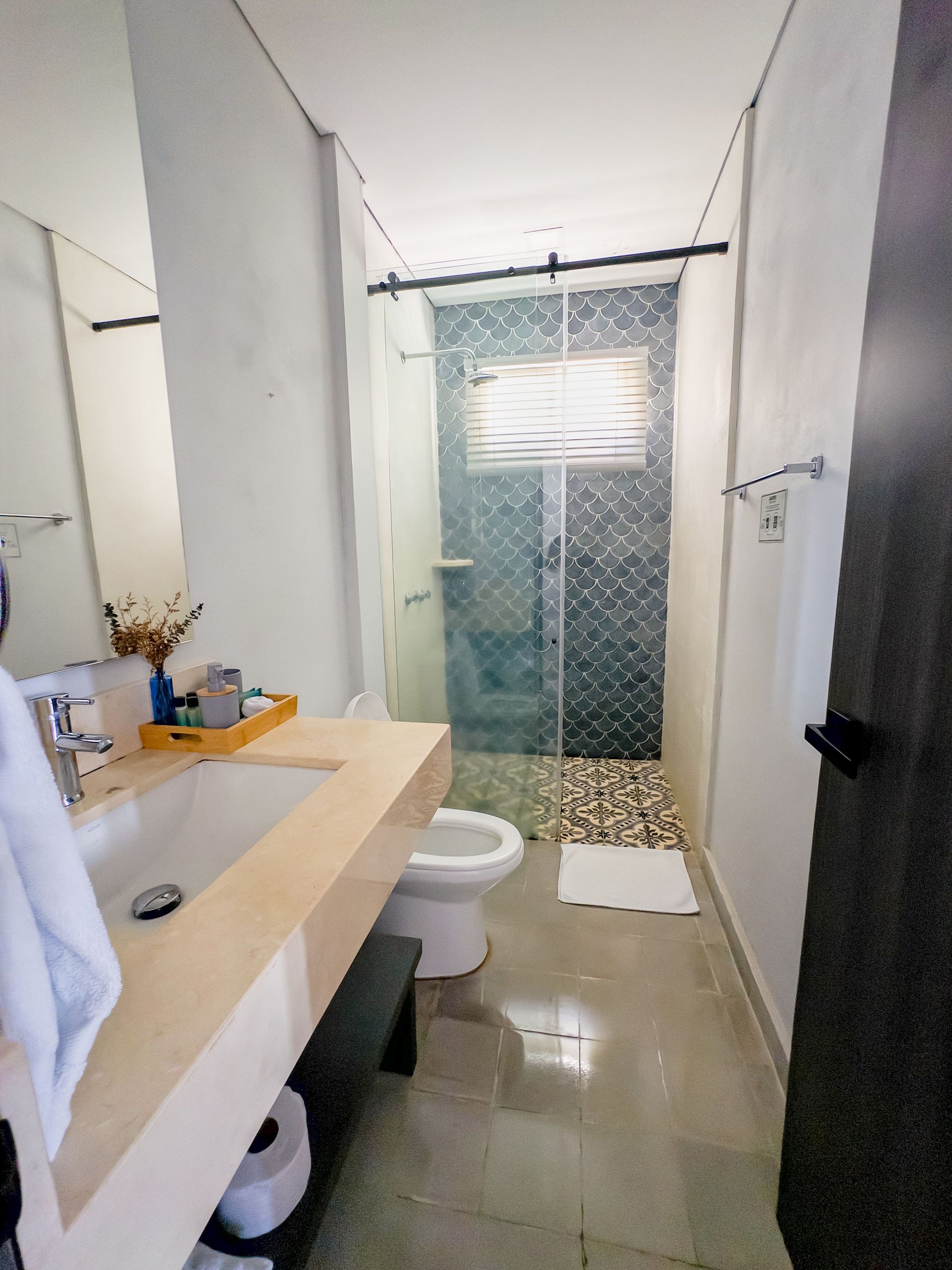 Baño de distribución alargada que dispone de lavabo, inodoro, ducha con puertas de cristal y encimera de piedra en color claro.