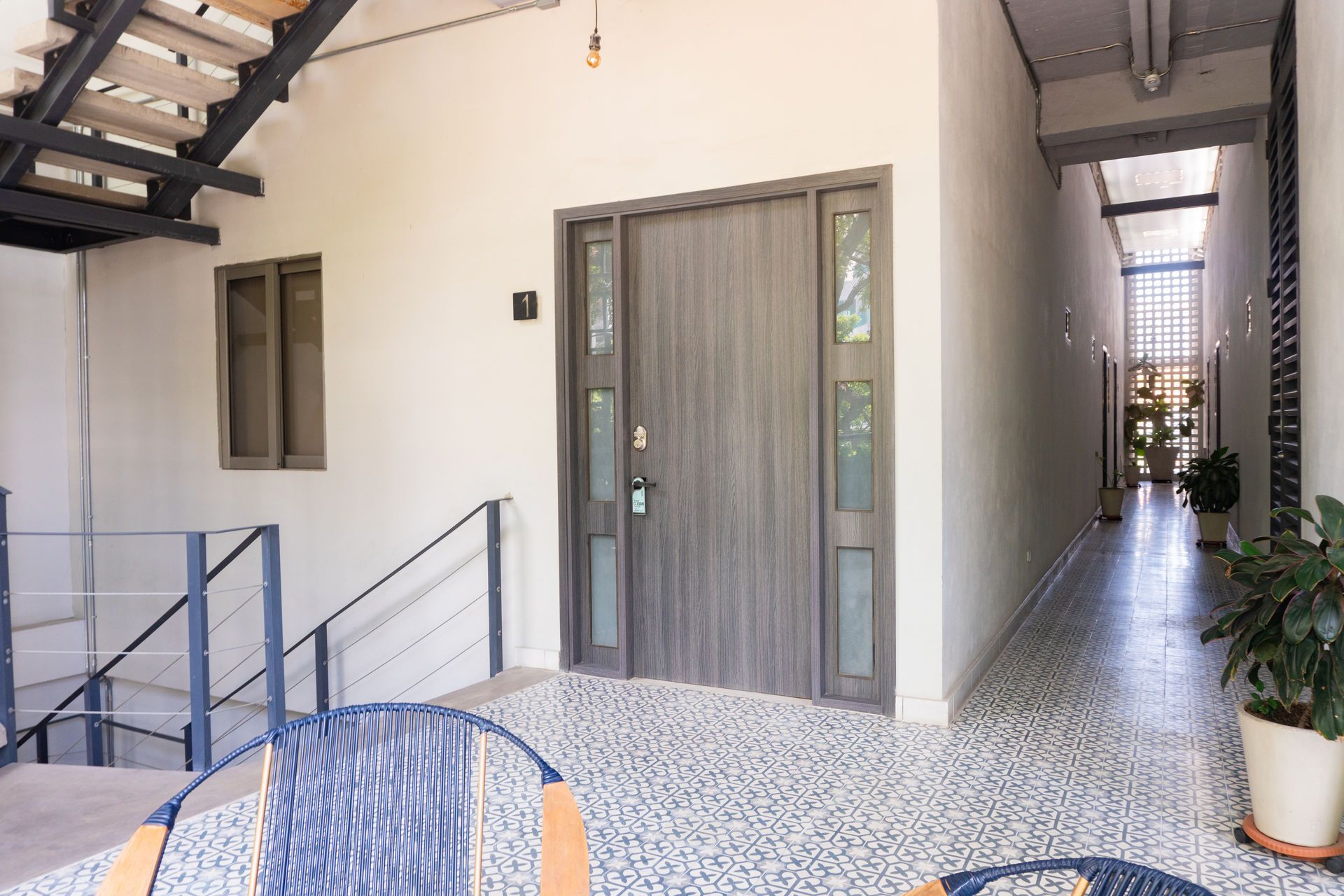 Entrada a un edificio con puerta gris, suelo de guijarros y escaleras. Un pasillo se extiende a la derecha.