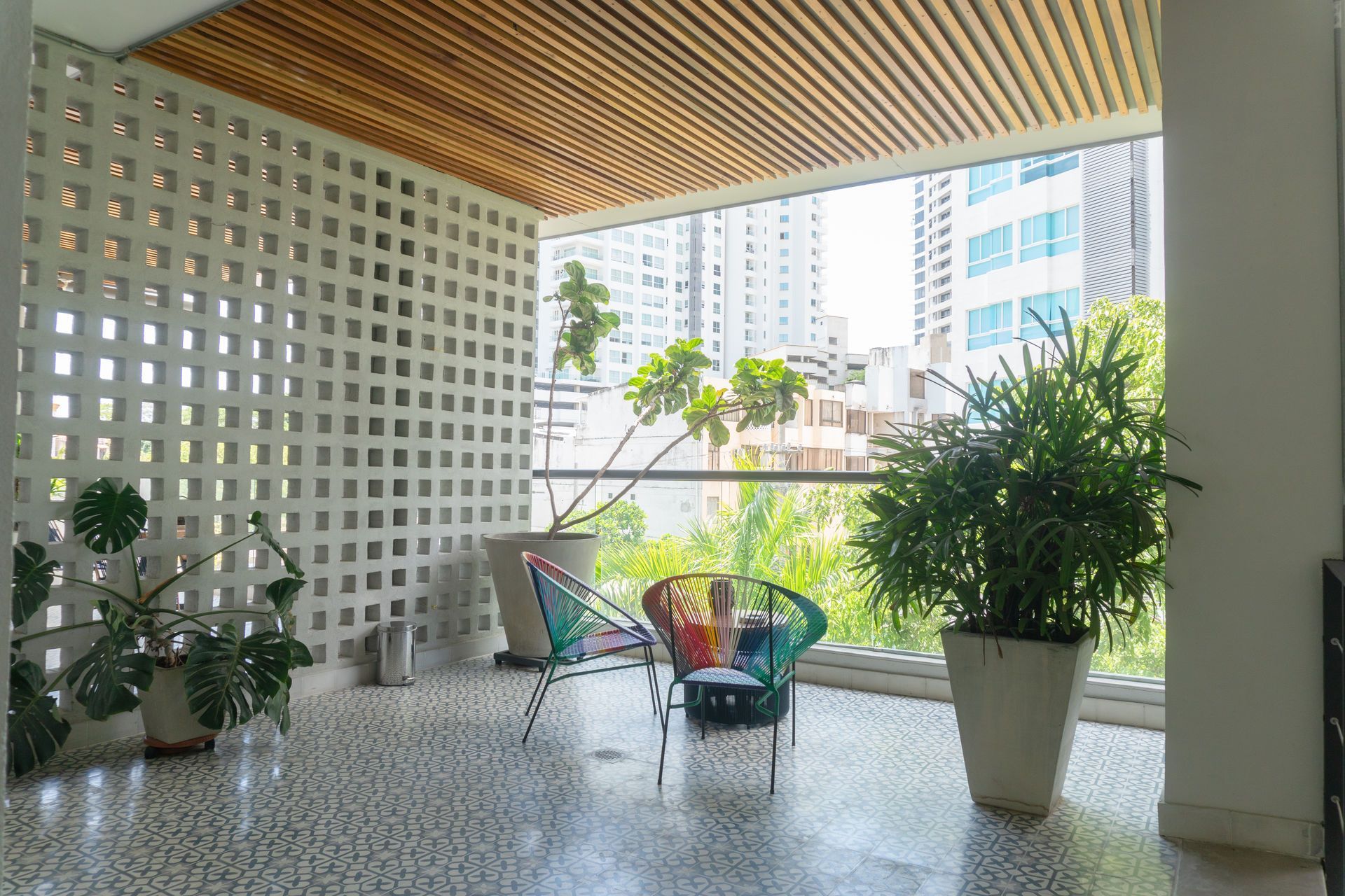 Balcón con piso de baldosas blancas, plantas en macetas, sillas de colores y vista a la ciudad.