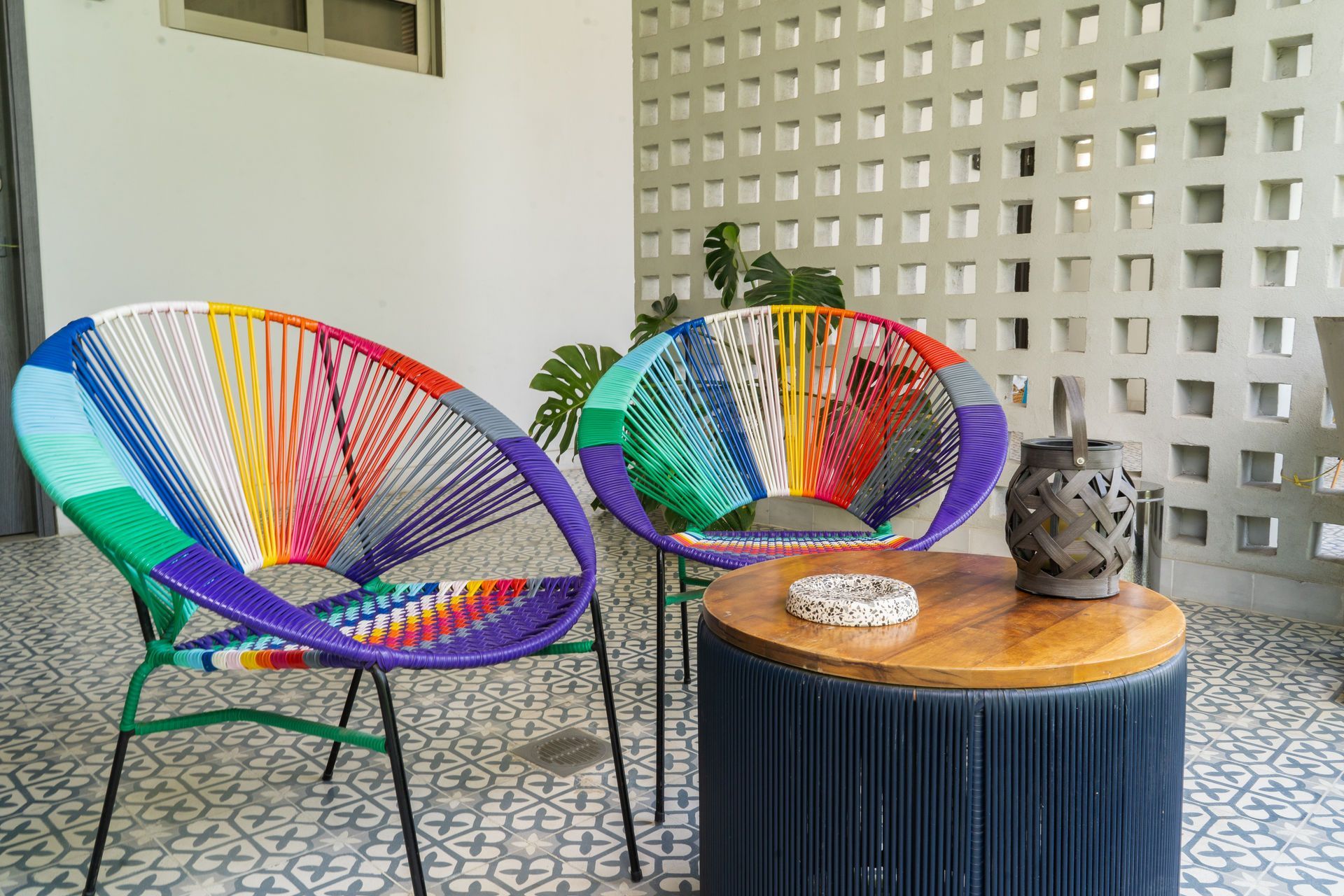 Dos sillas tejidas de colores en un patio estampado, con una mesa y una pared decorativa.