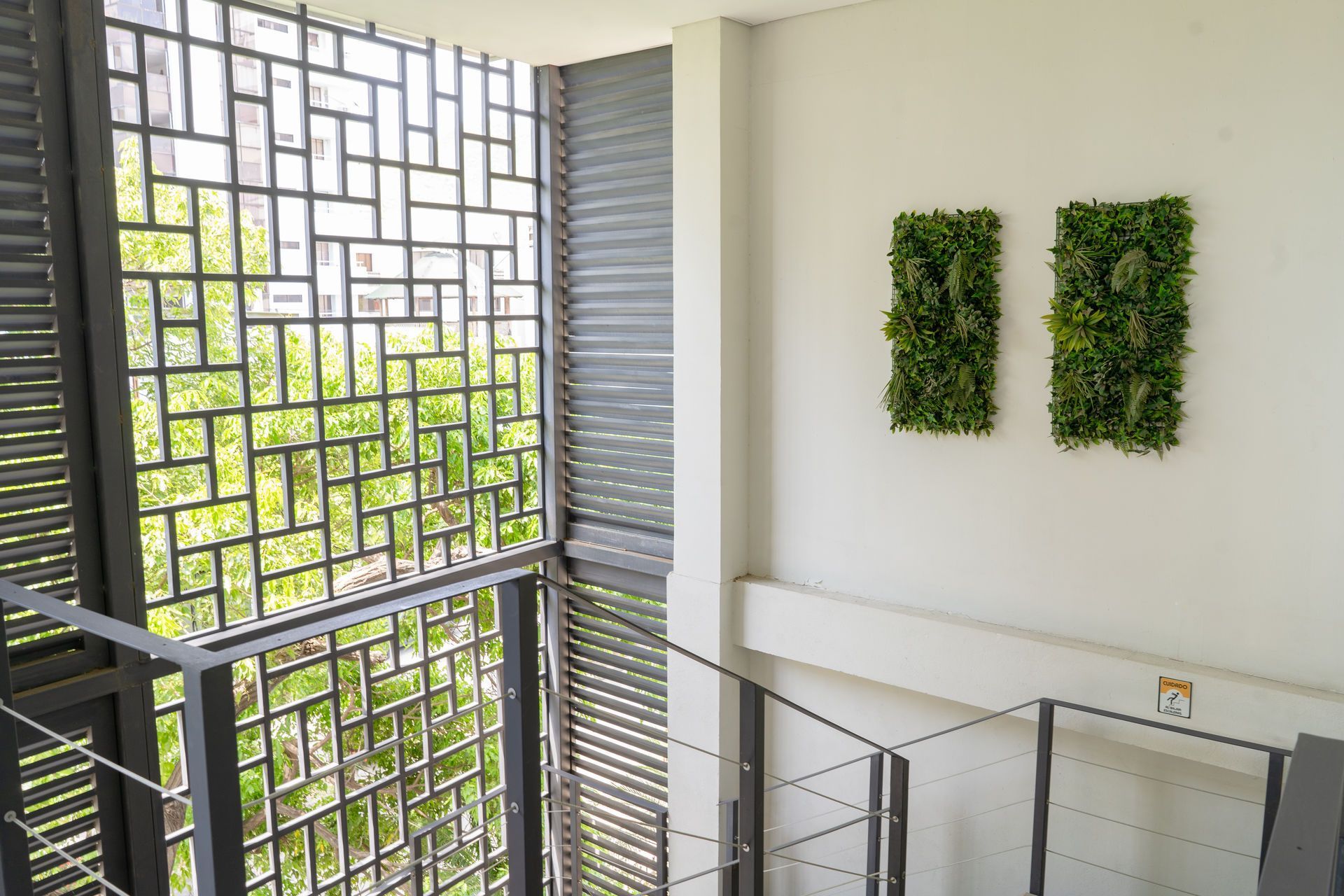 Escalera con barandilla metálica y paneles decorativos. Dos muros rectangulares de plantas verdes sobre pared blanca.