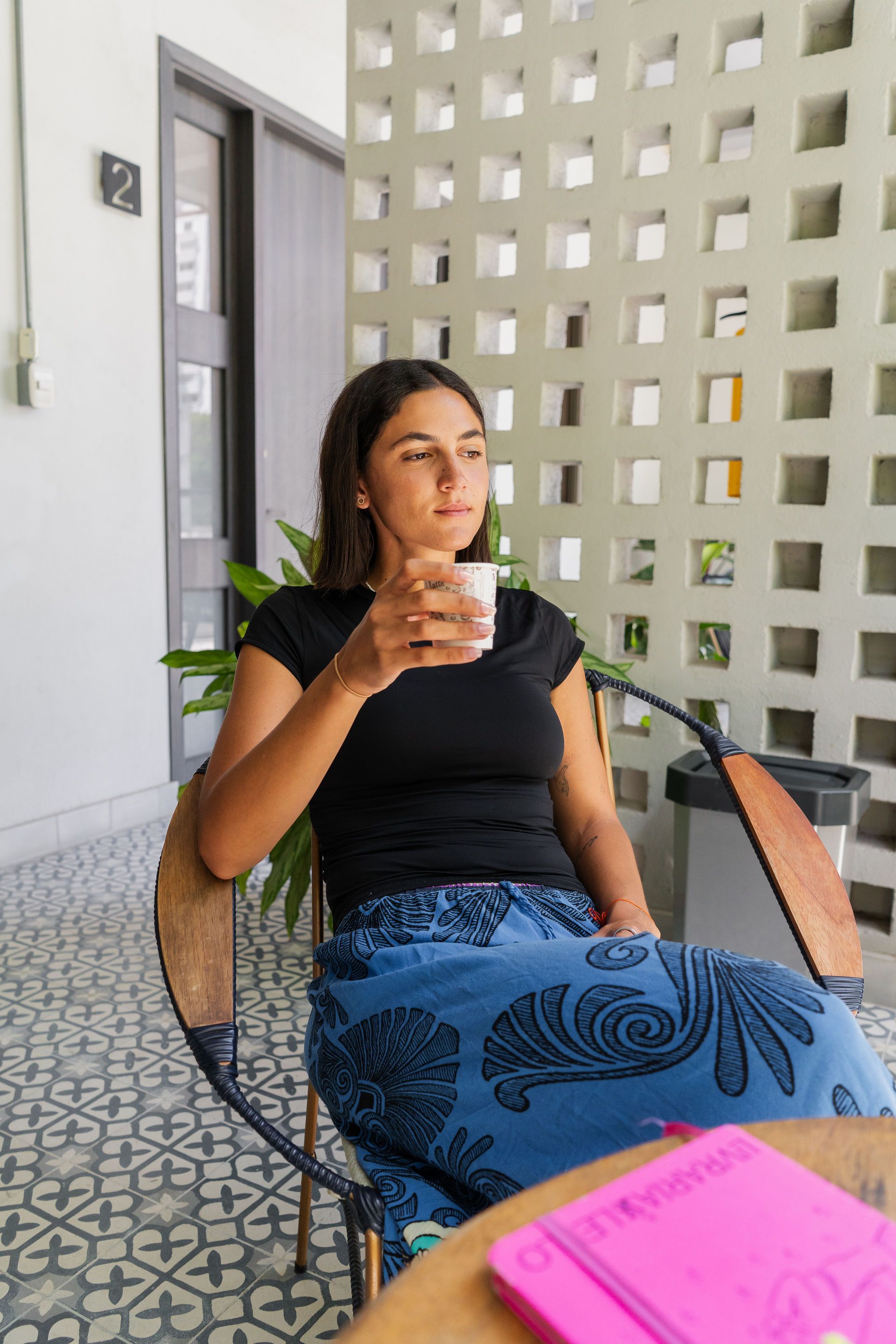 Mujer sentada, sosteniendo una taza, en un espacio exterior luminoso con paredes y suelos estampados, mirando hacia otro lado.
