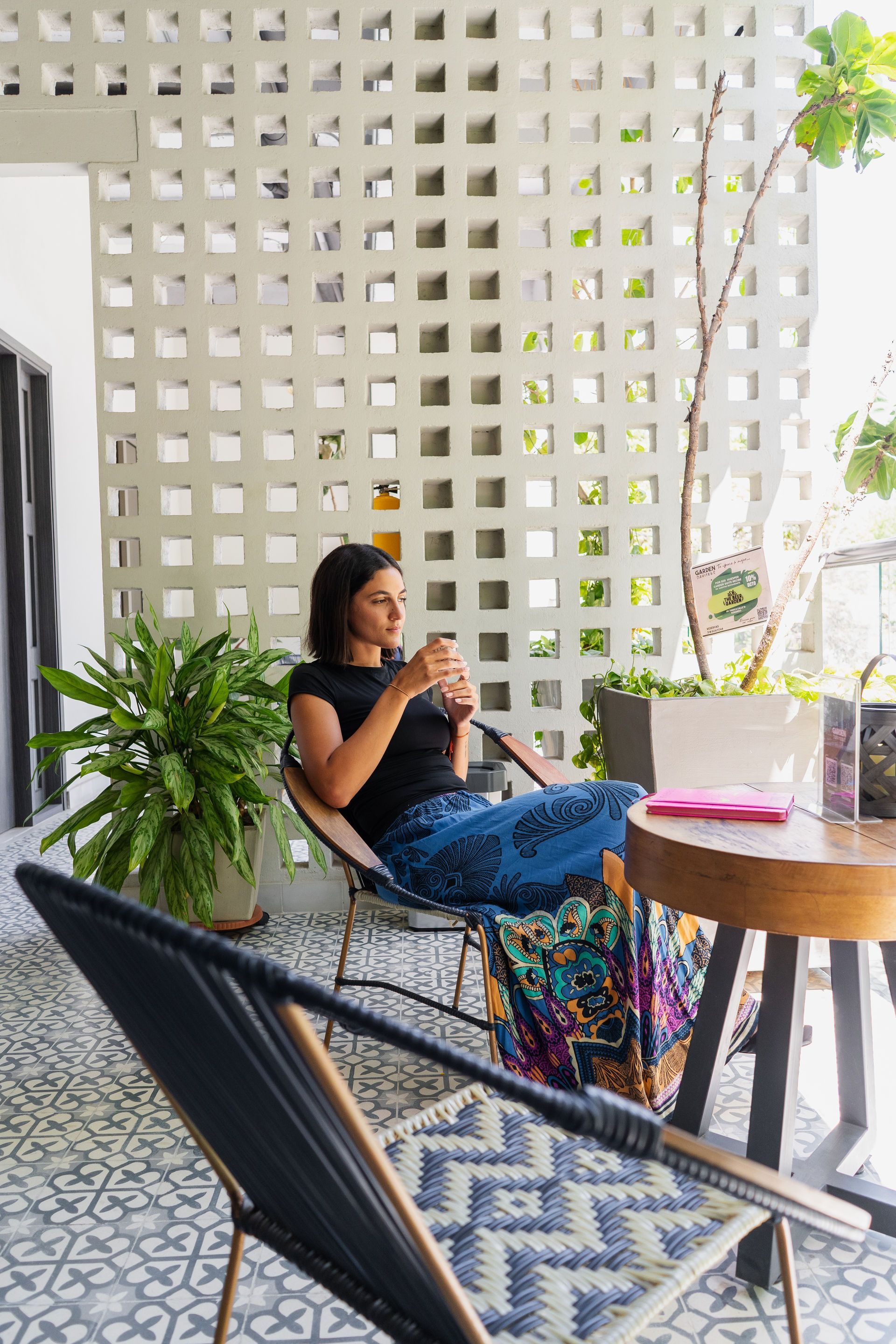 Mujer sentada en un balcón con una taza. Detrás de ella hay una pared blanca decorativa, plantas, suelo estampado y una mesa de madera.