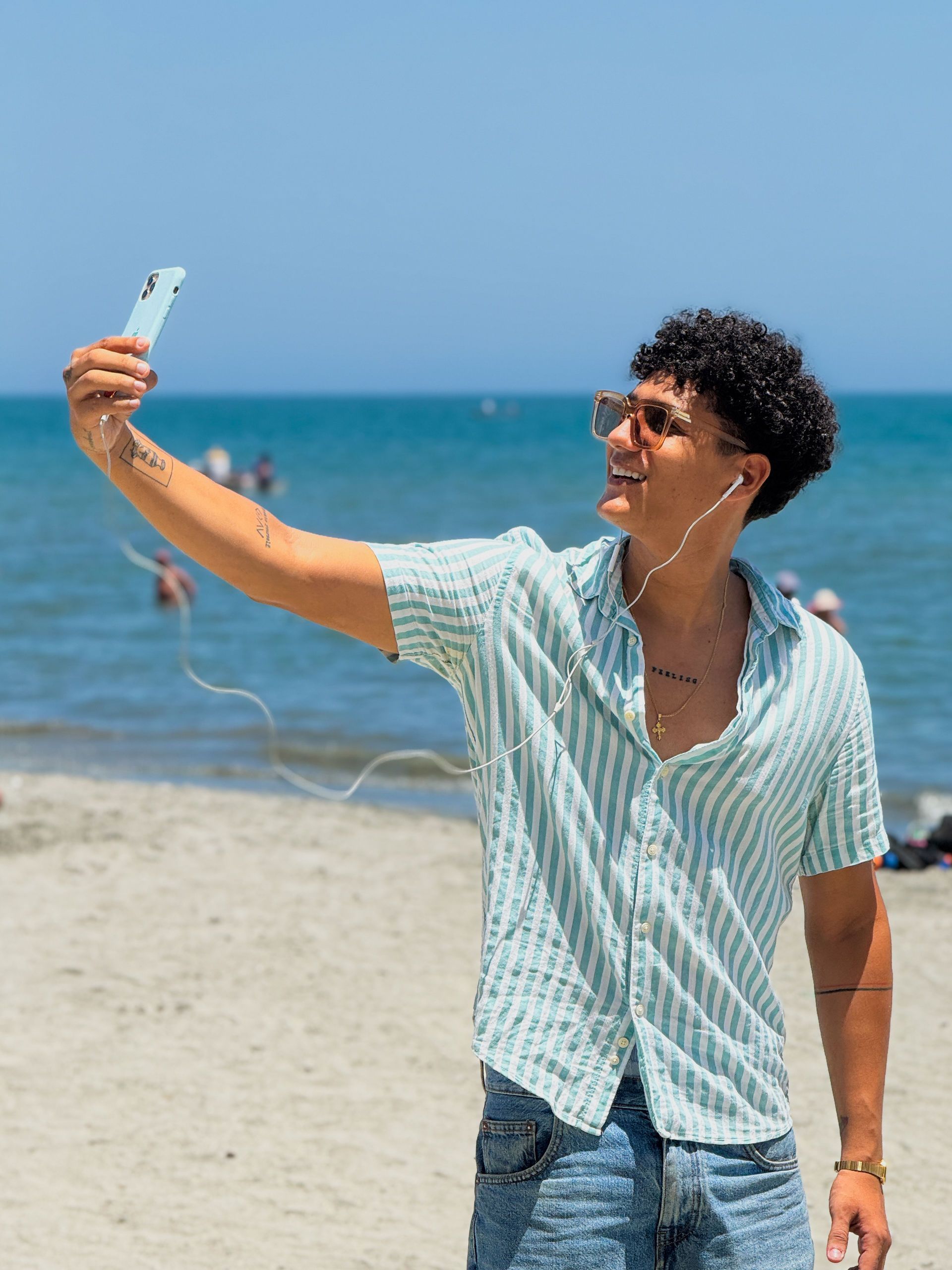 Hombre en la playa tomándose un selfie con un teléfono azul; camisa azul y gafas de sol. Océano de fondo.