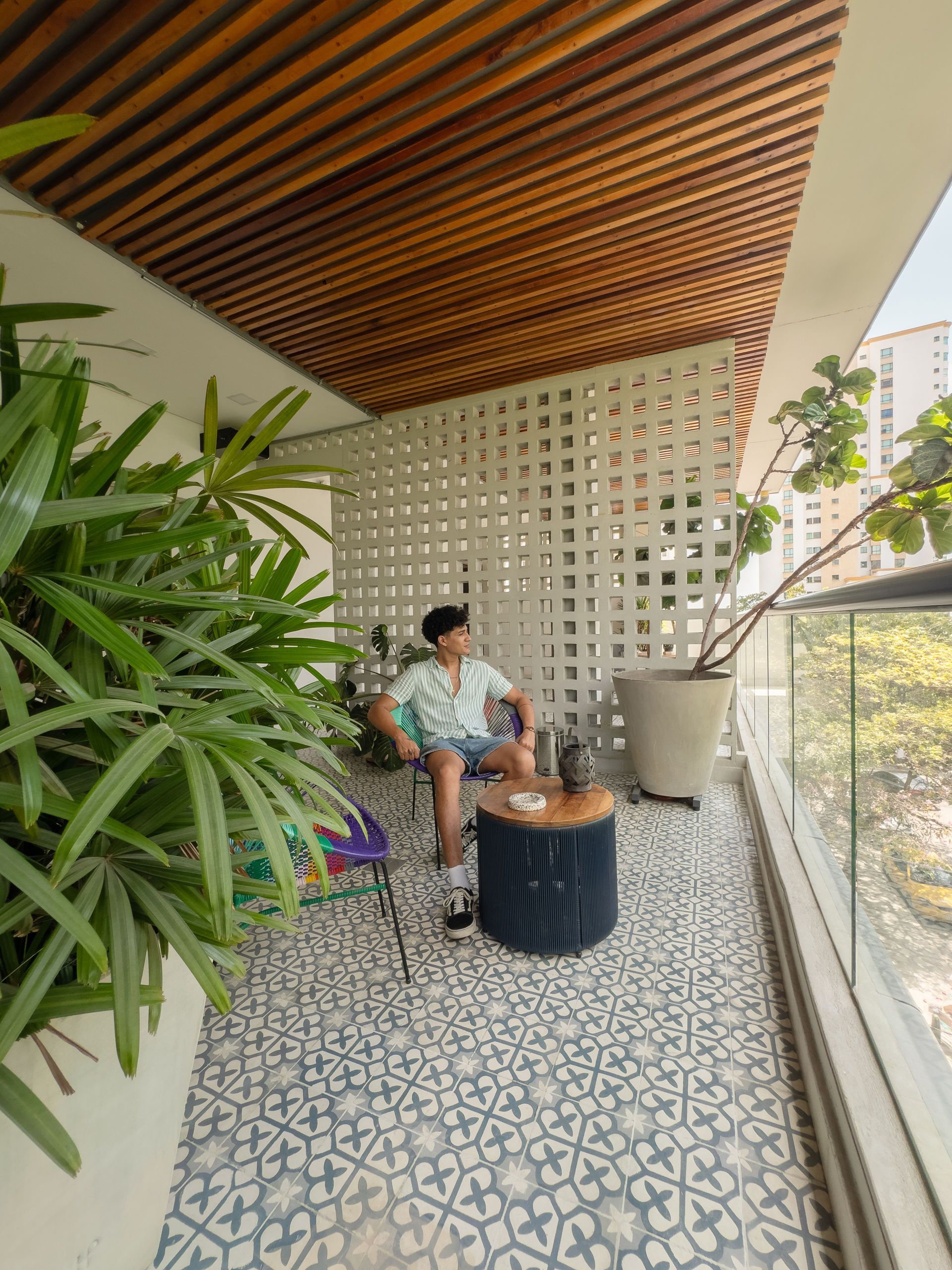 Hombre sentado en un balcón decorado con motivos florales, rodeado de plantas. Techo de madera, pared decorativa y barandilla de cristal.