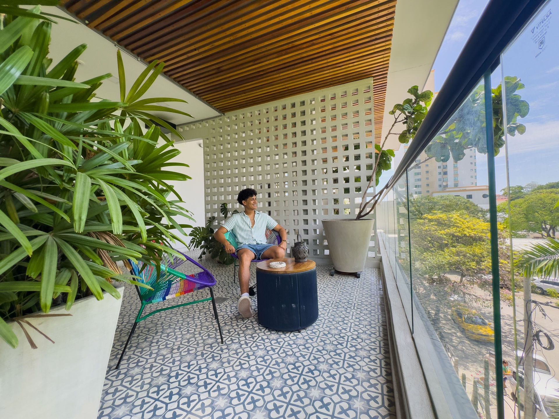 Hombre relajándose en un balcón con una silla colorida y plantas. Pared blanca estampada y vistas a la ciudad.