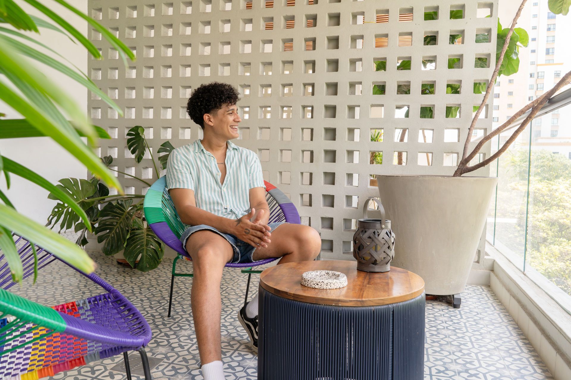 Persona en una silla colorida en un balcón, sonriendo, mirando hacia un lado, rodeada de plantas y una pared decorativa.