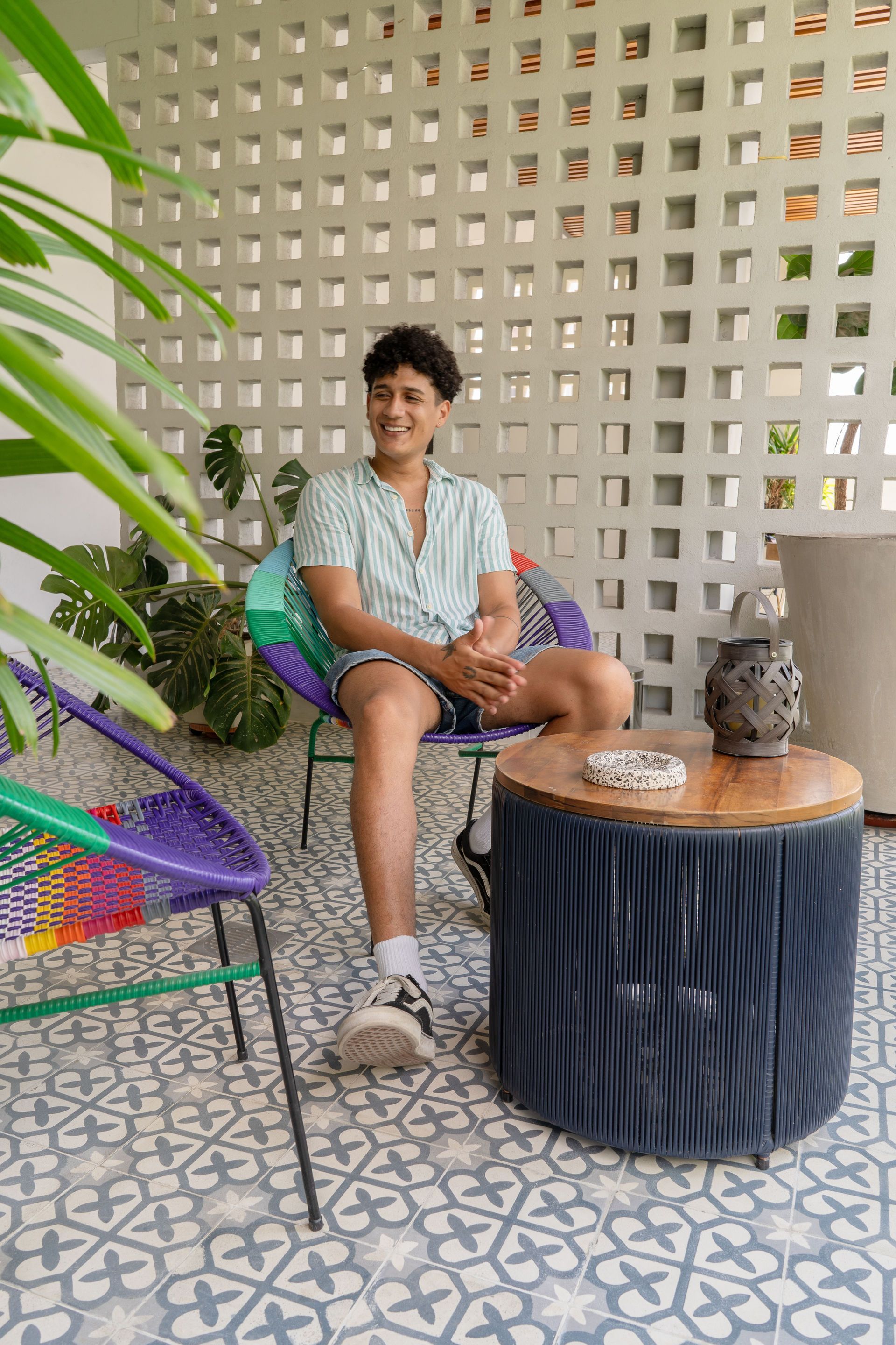 Un hombre con camisa estampada está sentado afuera, sonriendo. Junto a una mesa de madera y sillas de colores, frente a una pared de bloques blancos.