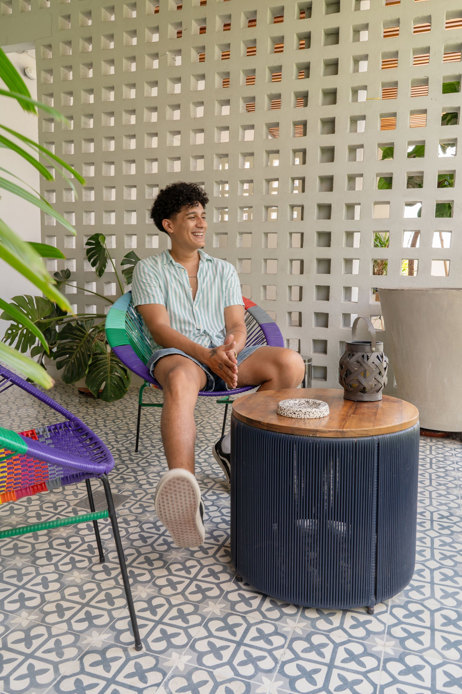 Hombre sentado en una silla colorida, sonriendo en un patio, con una pared estampada.