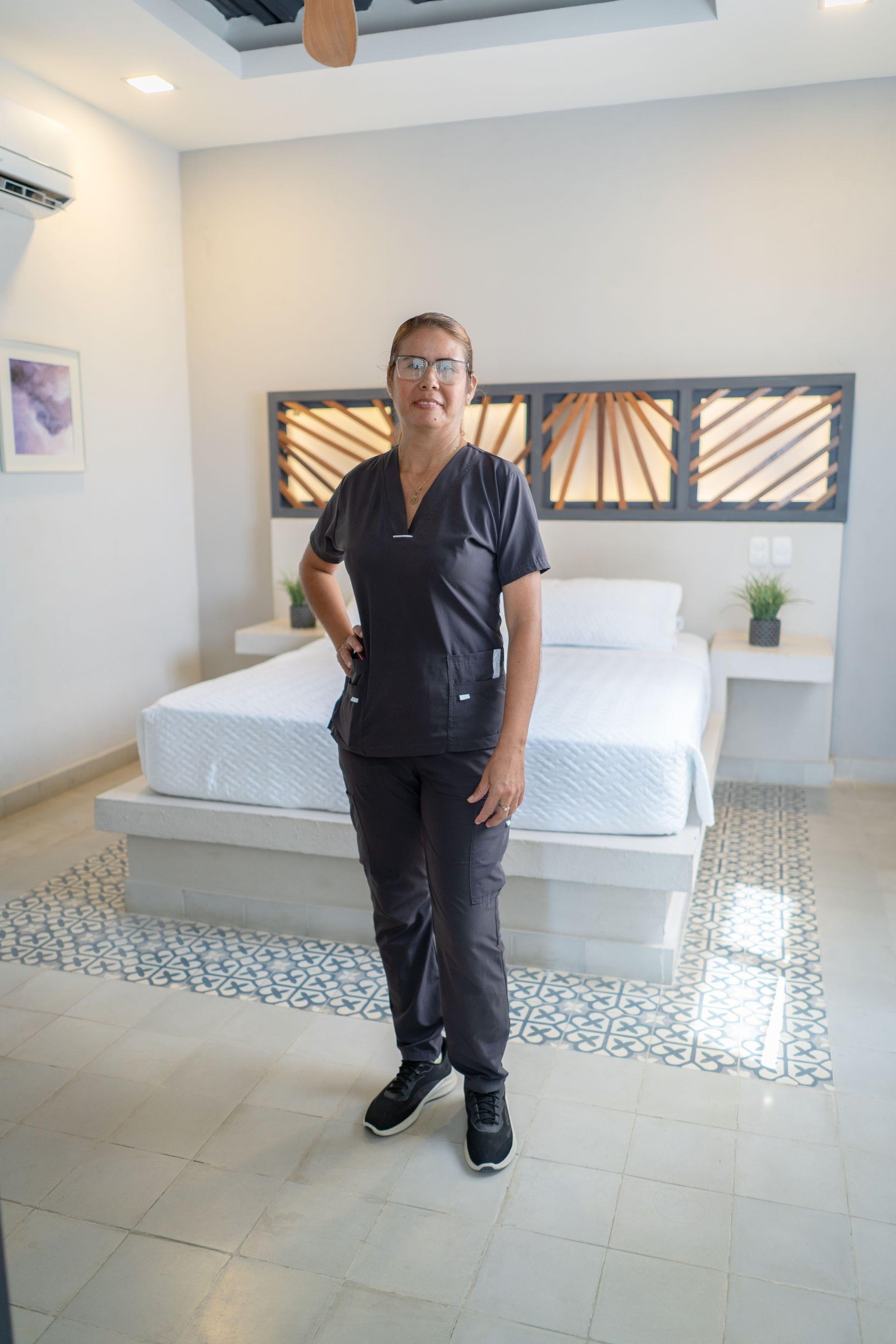 Una persona con uniforme médico oscuro está de pie frente a una cama. Interior de dormitorio con cabecero decorativo y suelo de baldosas.