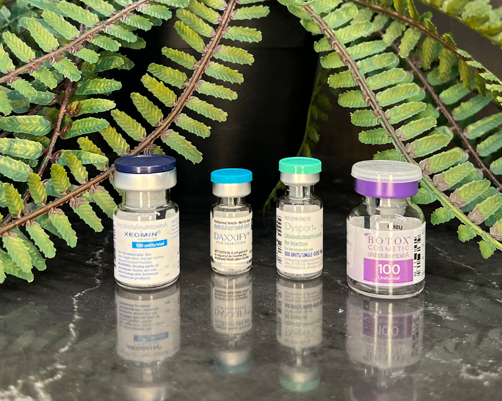 Four vials with different colored caps, sitting on a reflective surface in front of leafy foliage.