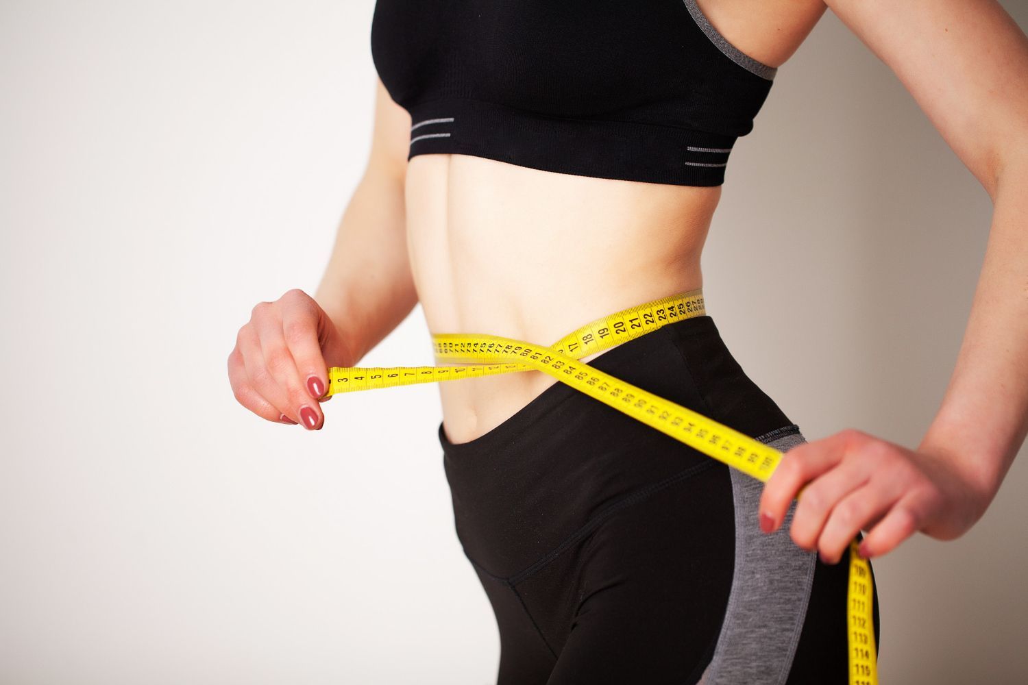 A person in athletic wear measuring their waist with a yellow tape measure against a plain background.