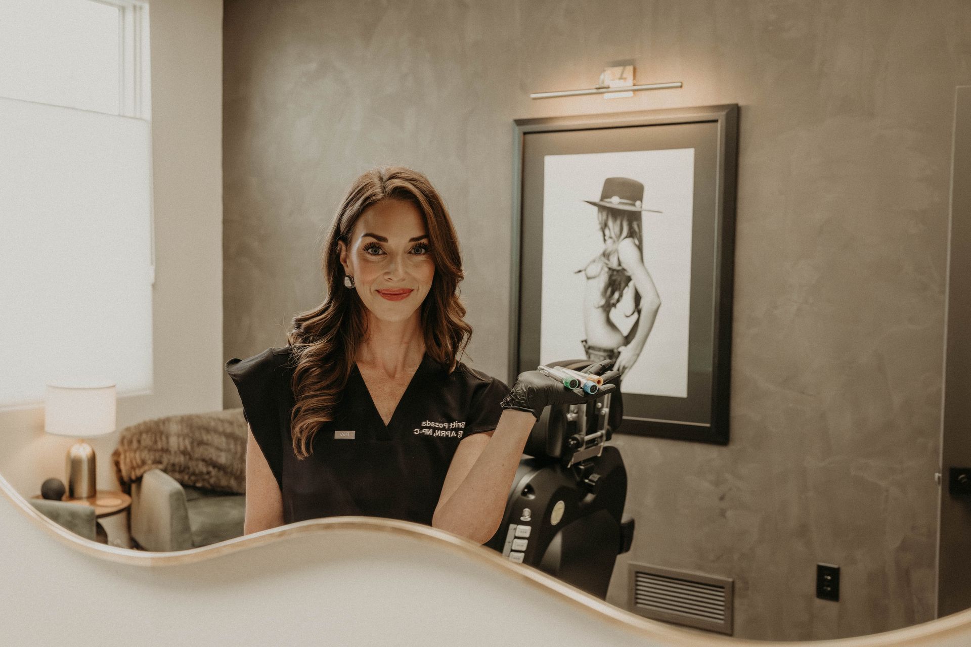 A woman in black scrubs poses in a modern office with a framed black-and-white fashion print on a textured gray wall.
