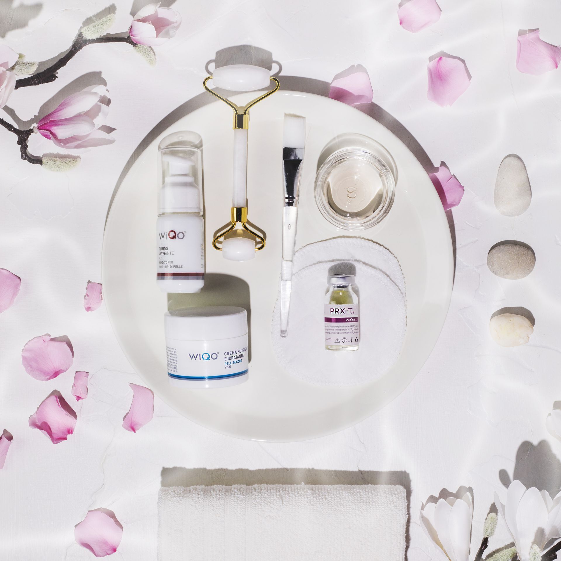 Skincare products arranged on a white surface with a jade roller, brush, and petals.