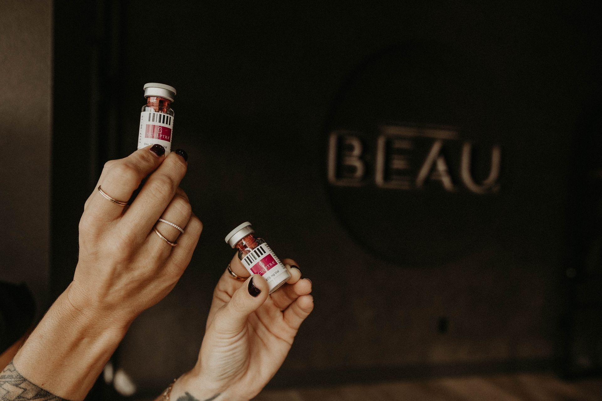 Hands holding two small red-and-white bottles in front of a dark sign reading “BEAU”